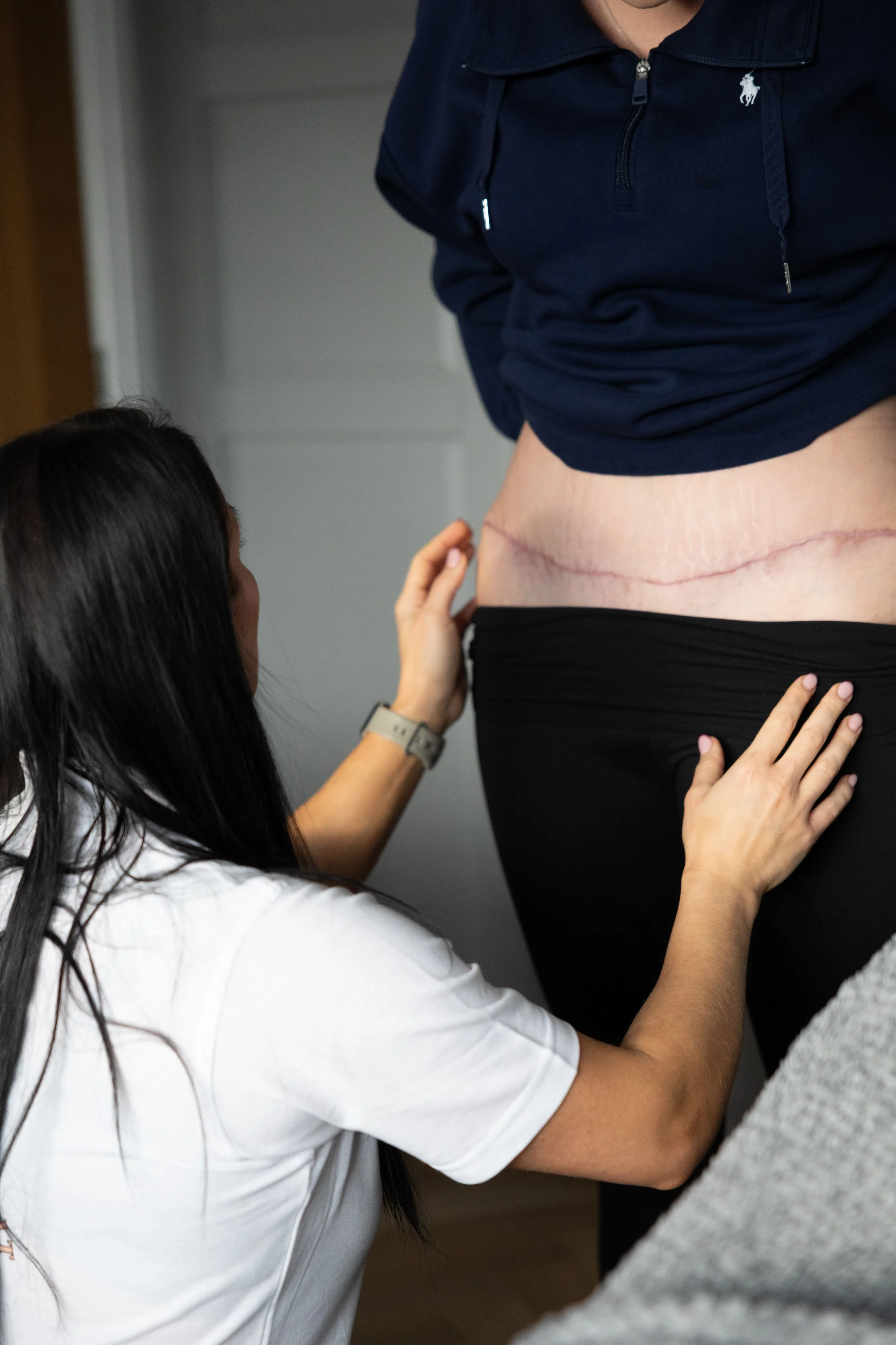 A woman with long dark hair in a white t-shirt kneeling and touching the lower back of a person with a surgical incision scar, wearing a dark blue hoodie and black pants.