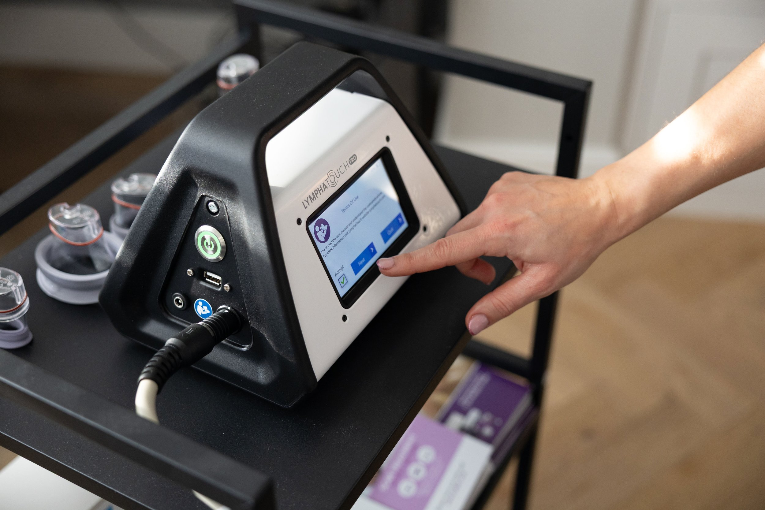 A person pressing a button on a medical device labeled 'LymphoTouch PRO' on a black cart with vials nearby.