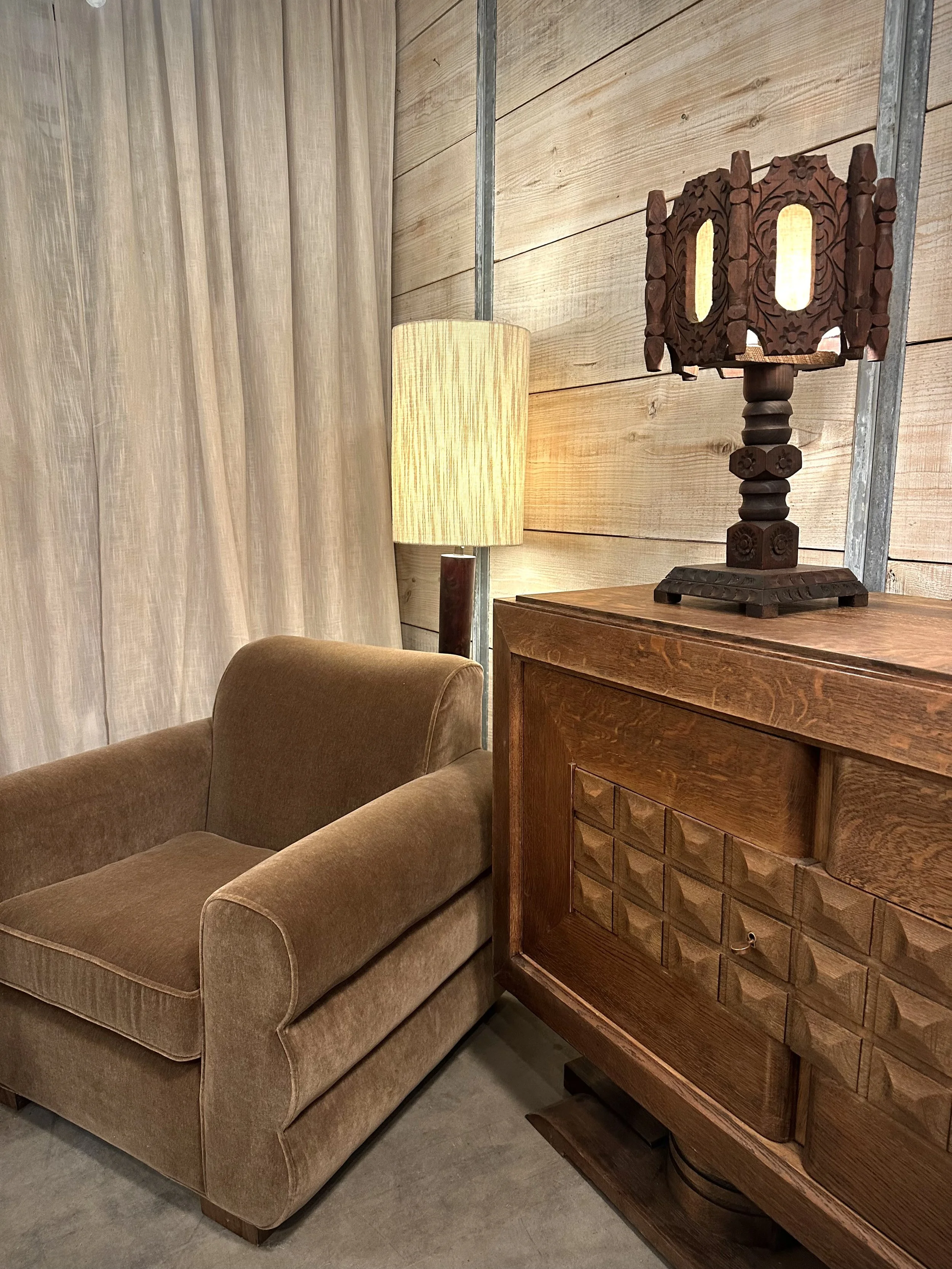 A living room corner with a brown upholstered armchair, a wooden sideboard, a beige cylindrical lamp, and a decorative wooden table lamp with intricate carvings on a wooden wall.