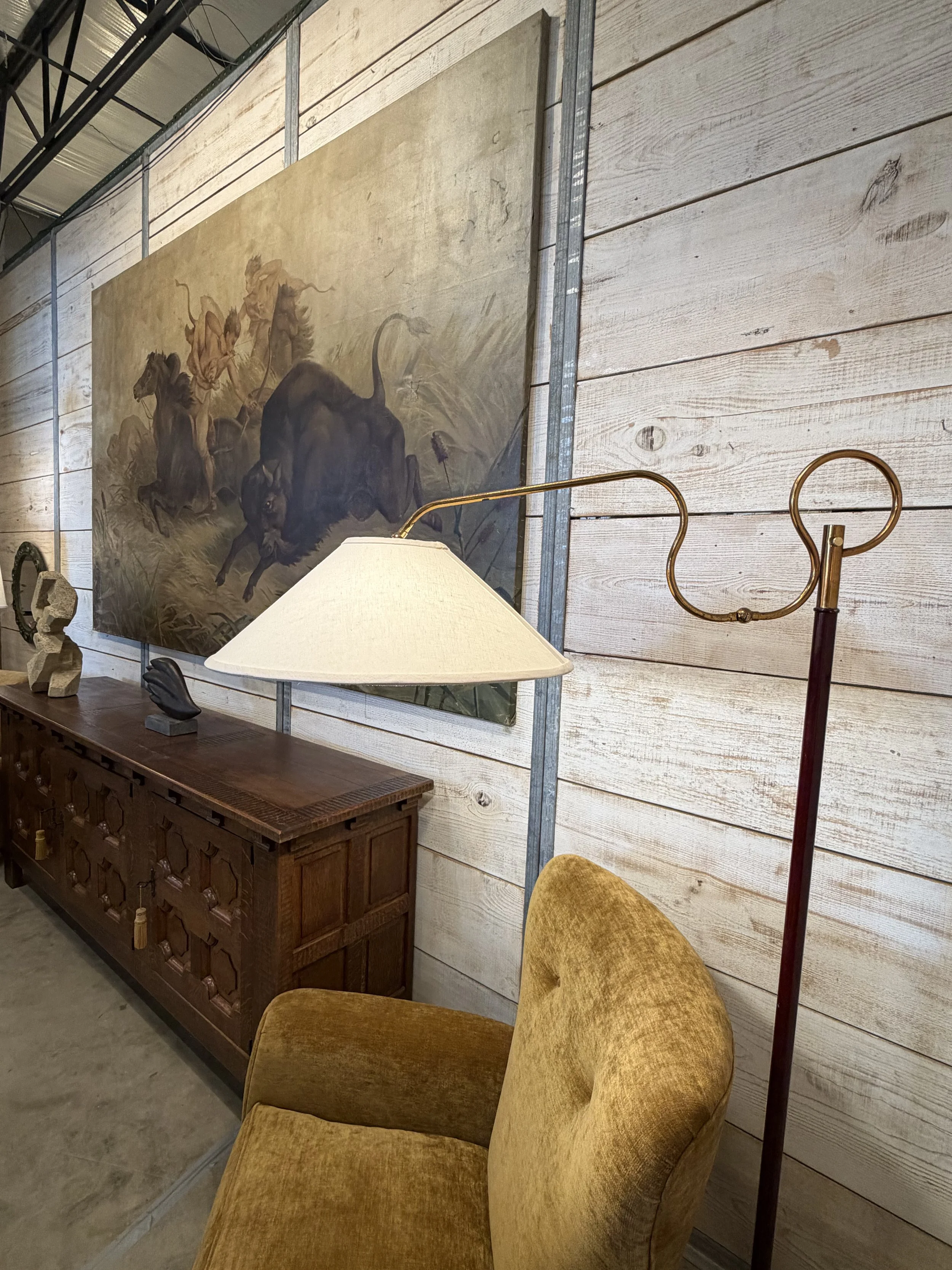 An interior scene with a vintage yellow armchair, a wooden sideboard, a lamp with a white shade, and a landscape painting of horses on a wooden wall.