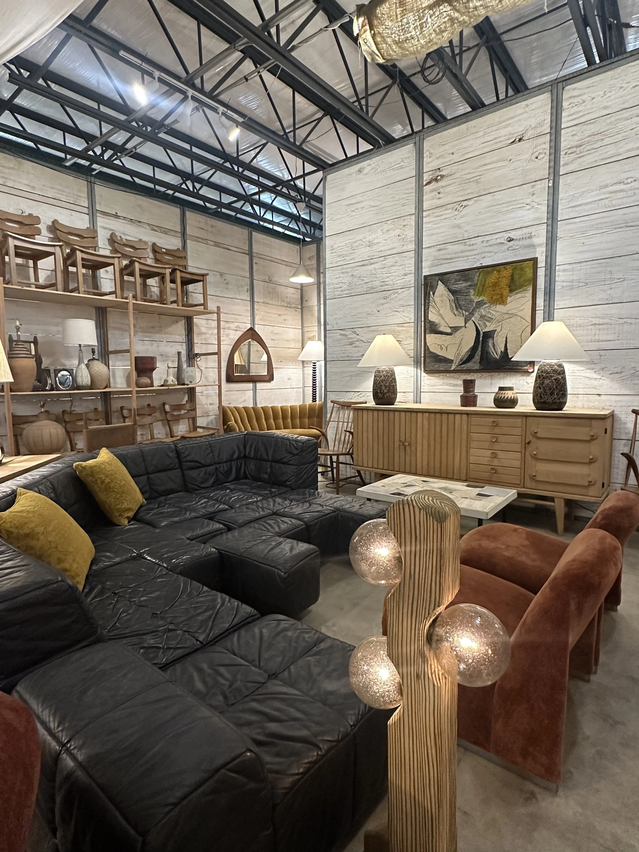 Living room with a black leather sectional sofa, yellow pillows, wooden chairs on a high shelf, a wooden sideboard with lamps and decorative pottery, a framed abstract artwork, and a floor lamp, with a ceiling featuring exposed metal beams and insula