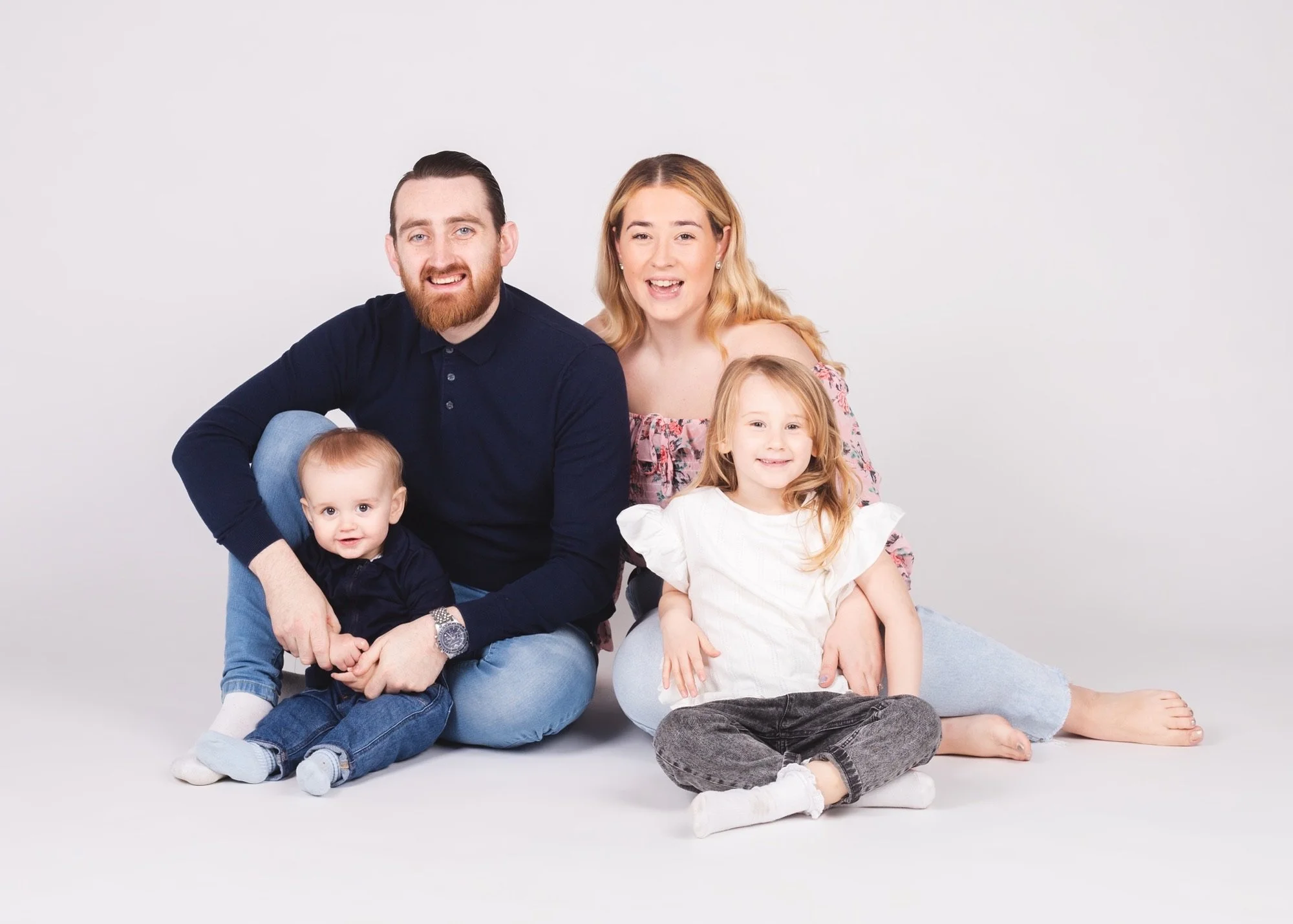 Family portrait of four people sitting on the floor against a white background. The man has dark hair and beard, wearing a dark shirt. The woman has blonde hair, wearing an off-shoulder floral top. The young girl has light brown hair in a ponytail, w