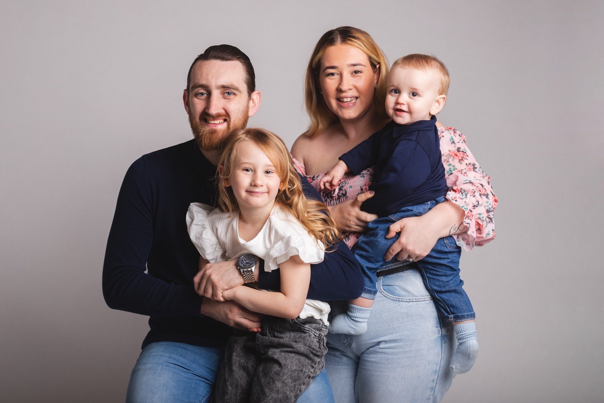 A happy family of four posing together in front of a plain grey background. The father with a beard and black shirt, the mother with blonde hair and floral top, a young girl with red hair and a white top, and a toddler boy in navy and jeans.