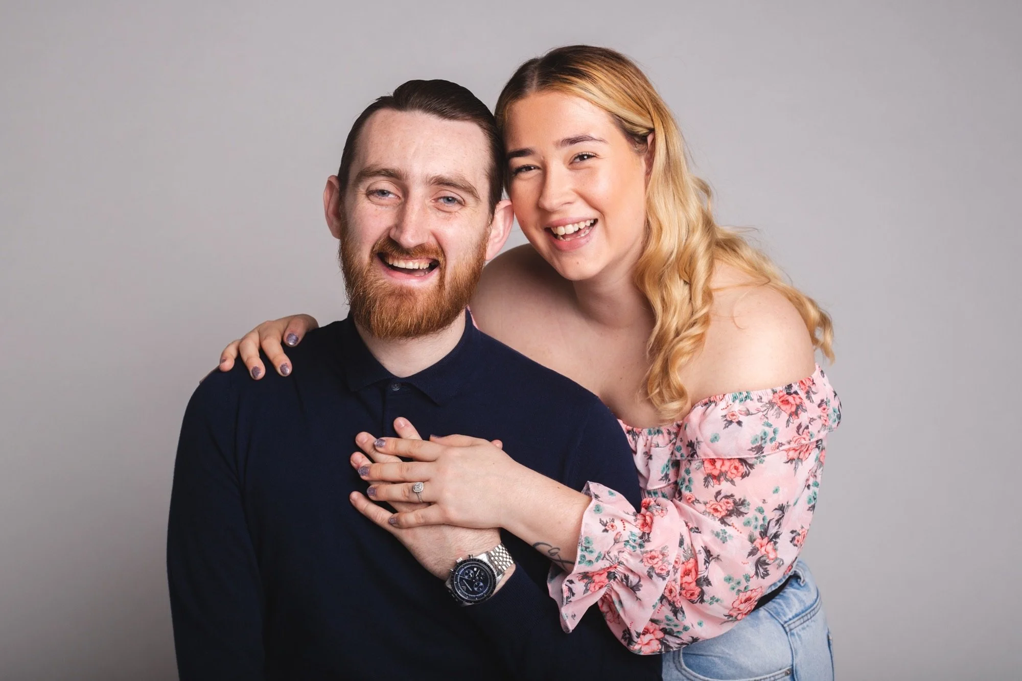 A happy couple posing together, with the woman standing behind and hugging the man, both smiling and looking at the camera, against a plain gray background.