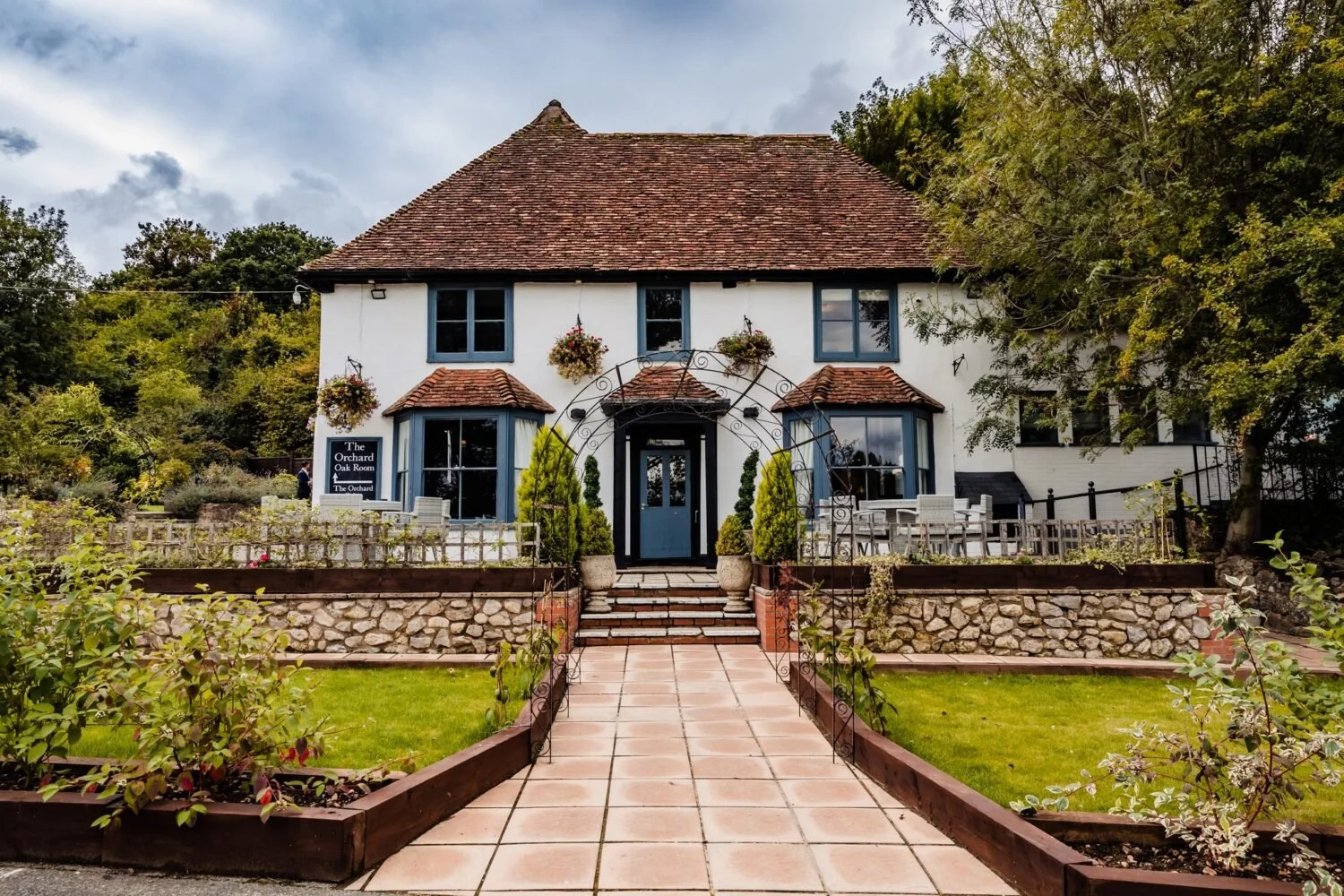 Charming two-story white house with blue window frames, a red tiled roof, and a garden front yard with a pathway, stairs, and potted plants.