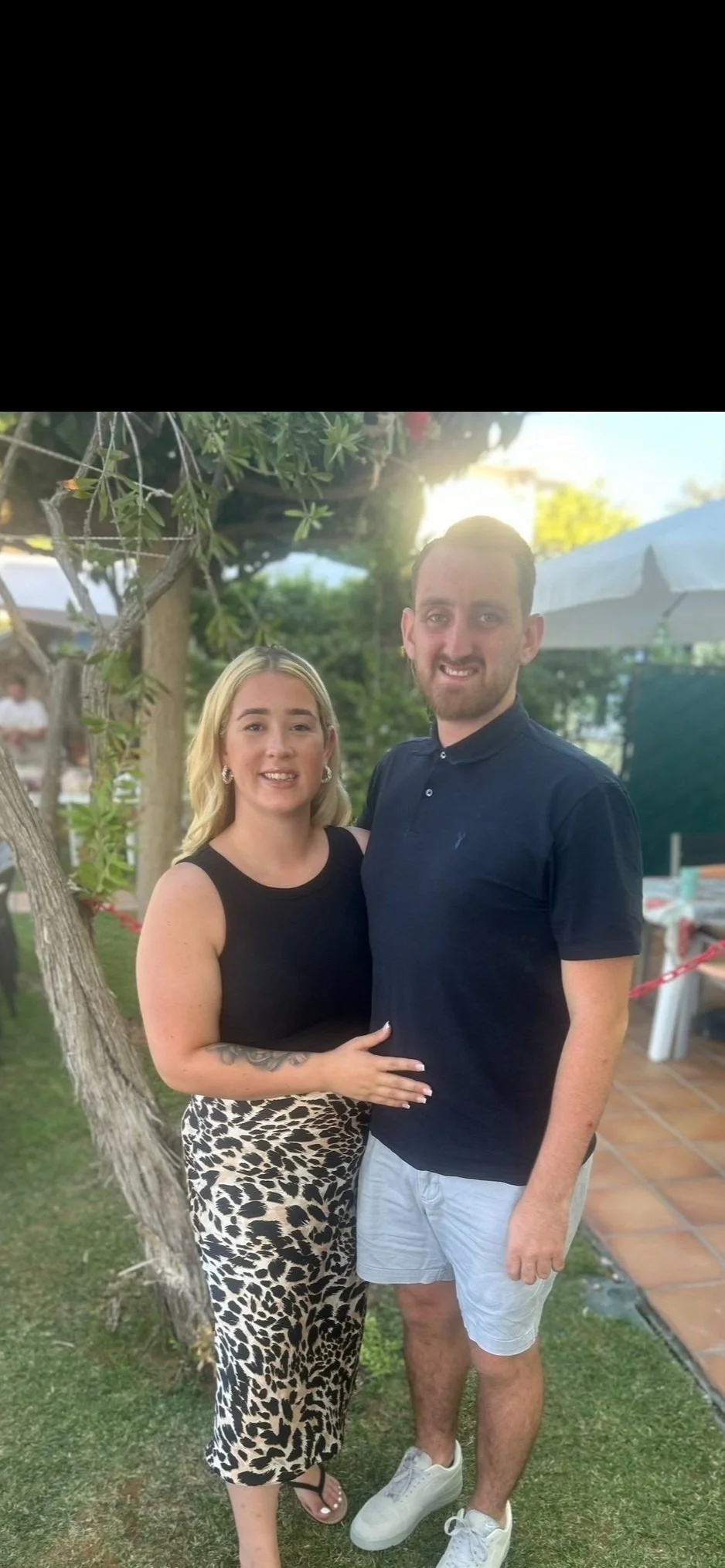 A woman and a man standing outdoors, smiling, with greenery and a tree in the background, at sunset.