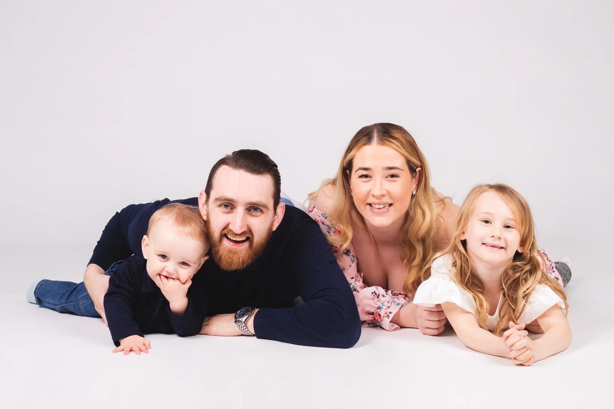 A family of four lying on the white floor together, smiling at the camera, with a plain white background.
