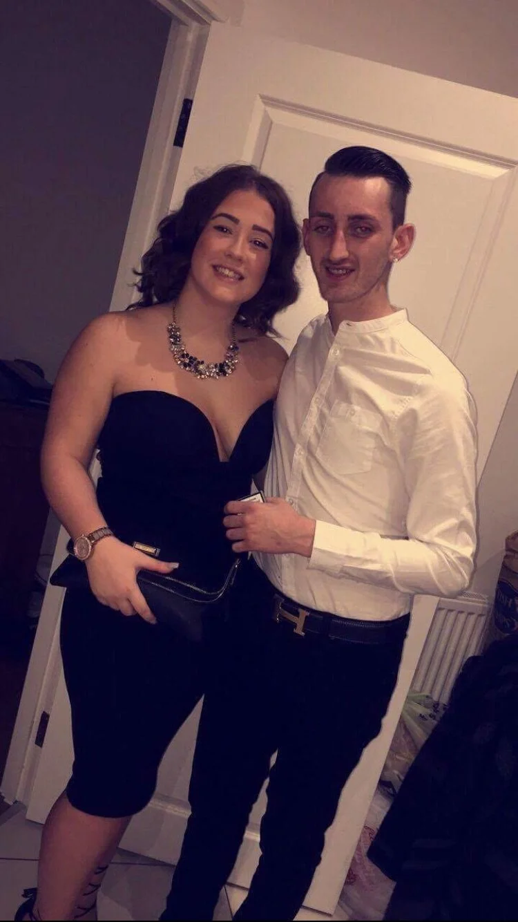 A young woman in a strapless black dress with a statement necklace and a man in a white shirt and black pants, standing together indoors and smiling.