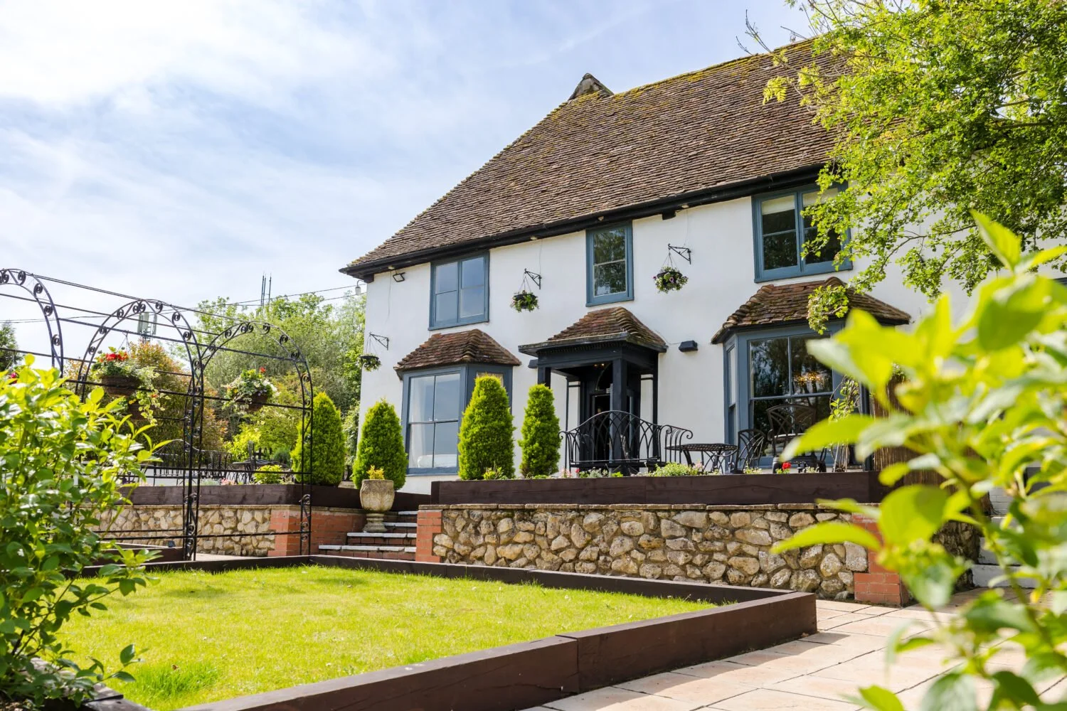 White two-story house with blue window frames and a brown tiled roof, surrounded by lush green trees and plants, with a patio and garden area featuring black wrought-iron furniture and decorative arches.