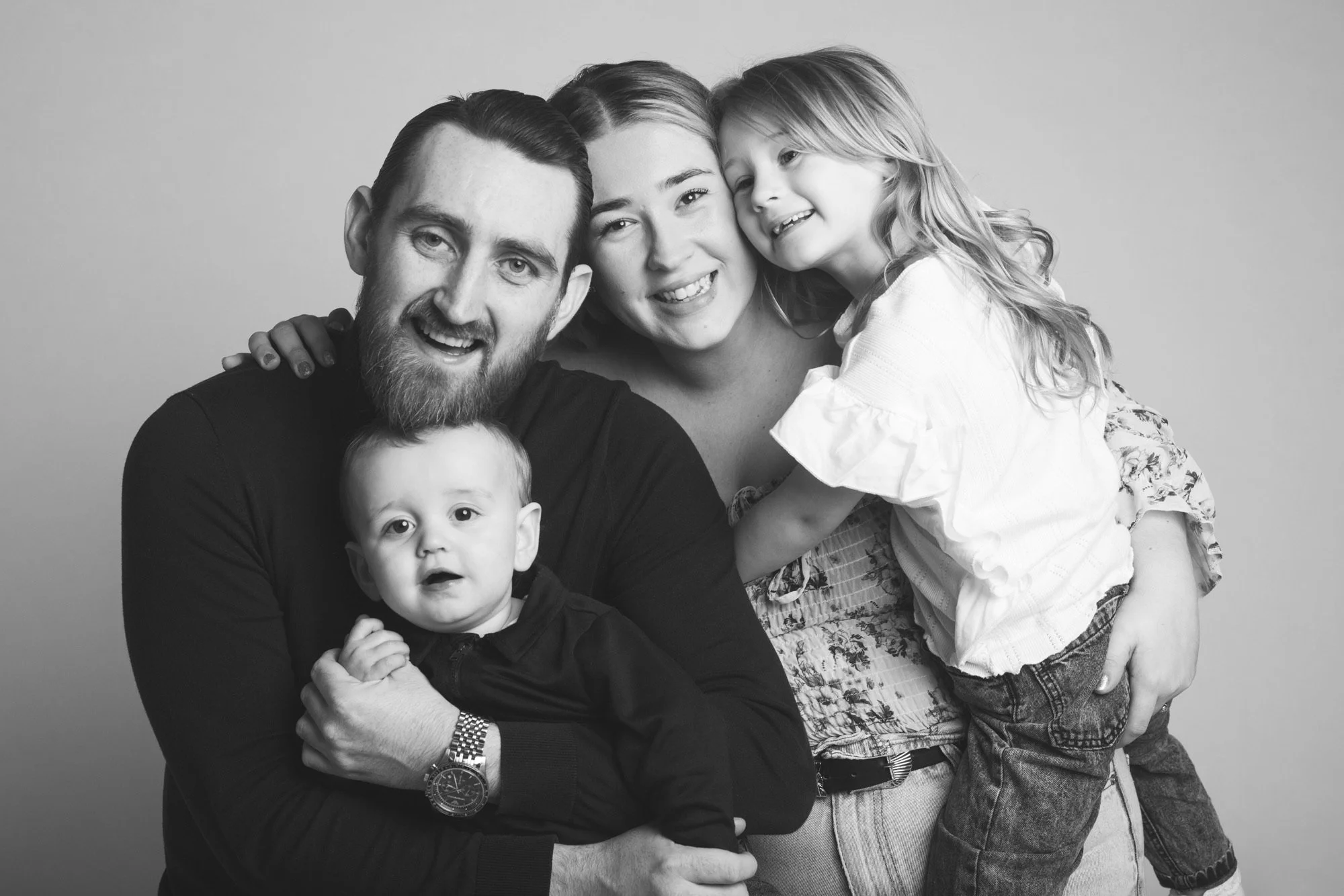 Black and white photo of a happy family of four smiling and hugging together, consisting of a man, a woman, a young girl, and a toddler boy.
