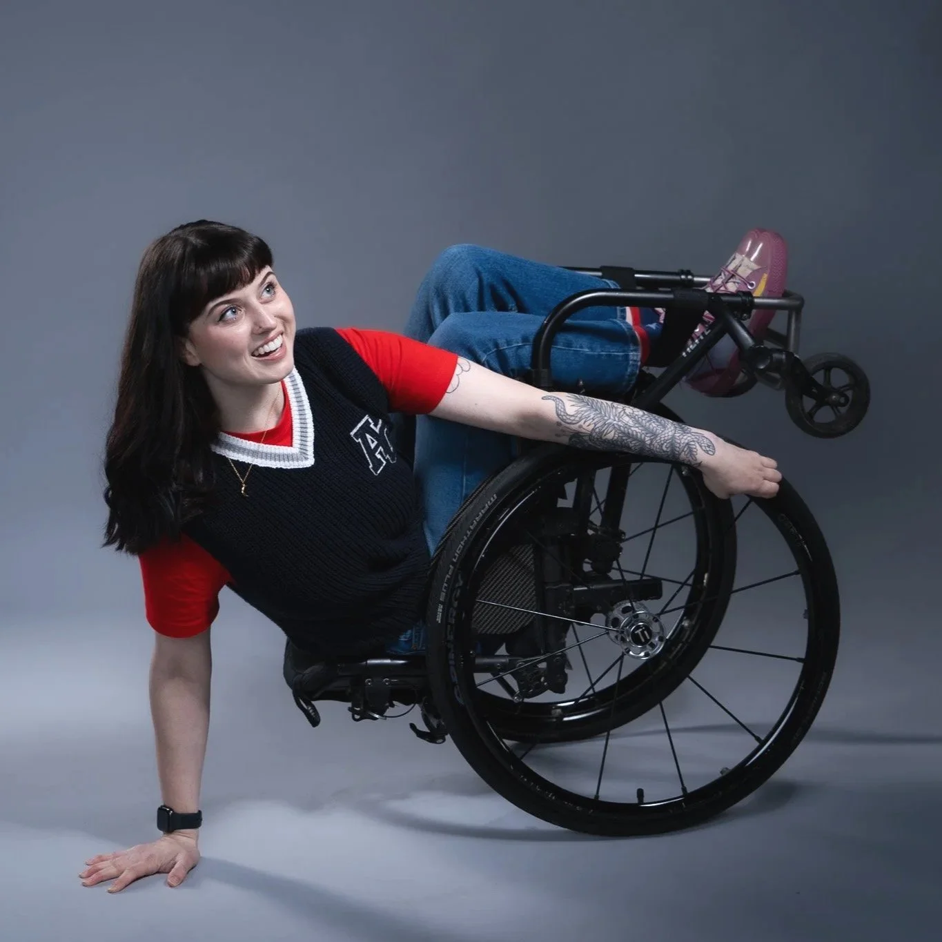 A young woman with dark hair, smiling, in a wheelchair, leaning on one hand on the ground with the other arm extended, wearing a black vest over a red shirt, jeans, and pink shoes, against a plain gray background.