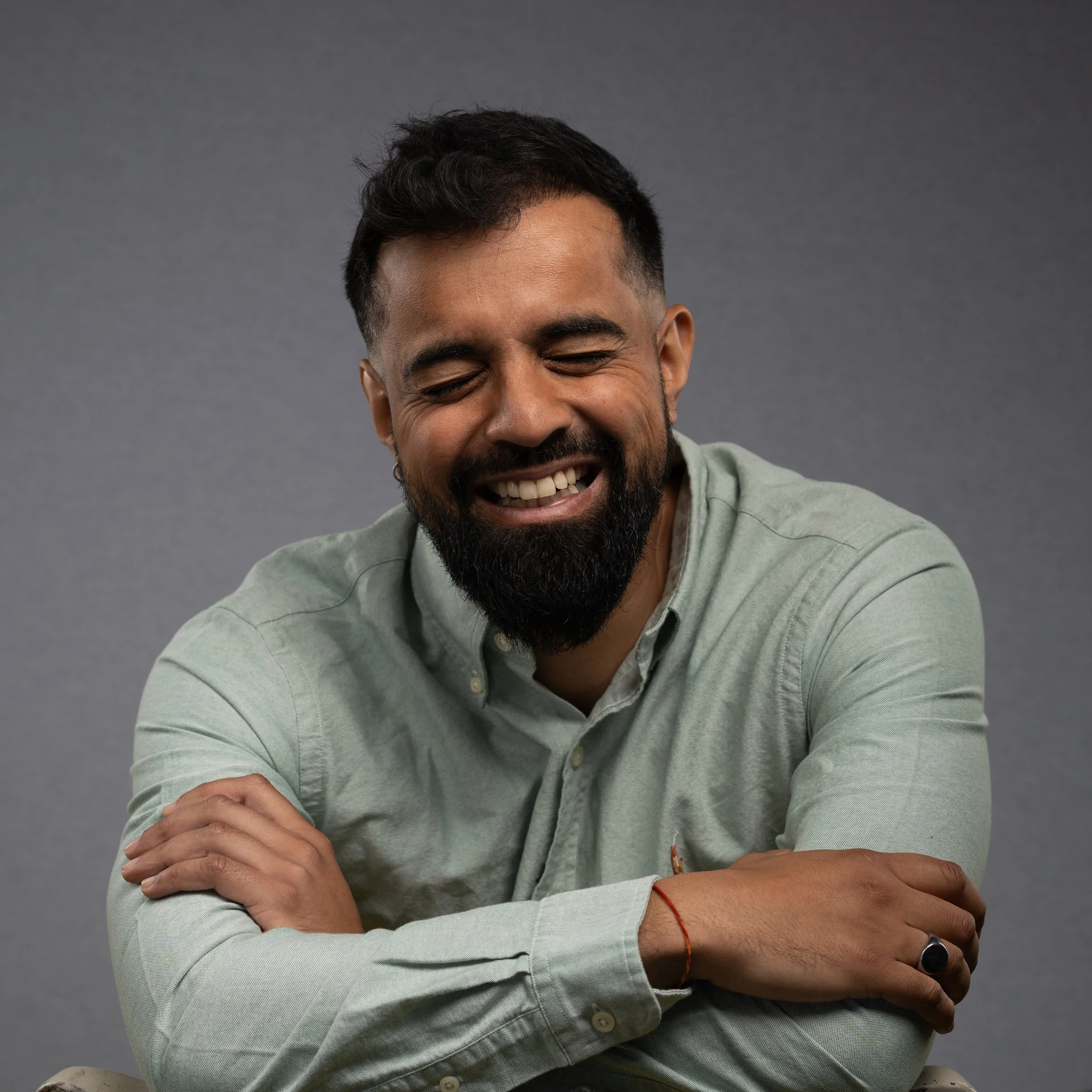 A man with a beard and short dark hair smiling with eyes closed, wearing a light green button-up shirt, sitting with arms crossed against a gray background.