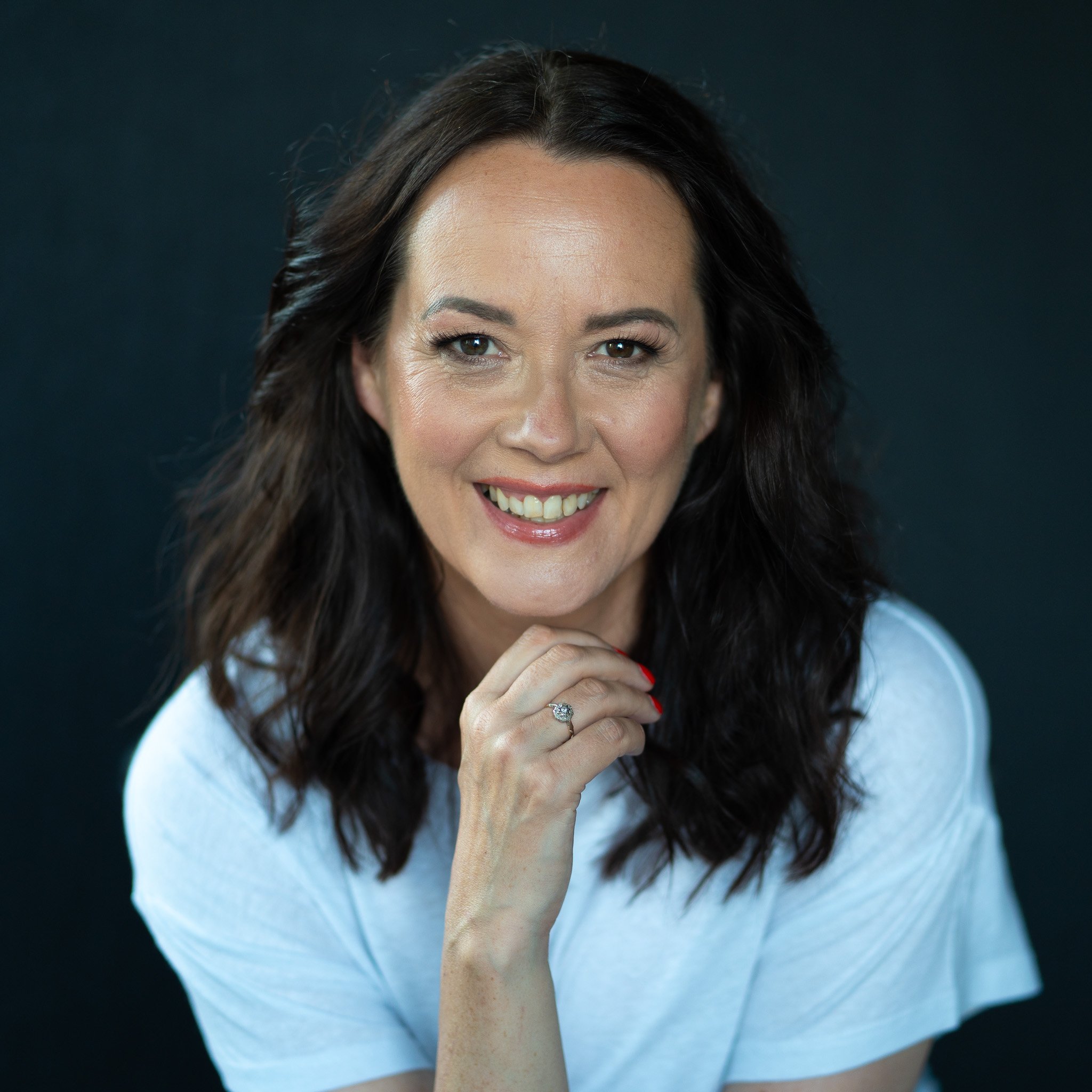 Close-up portrait of a smiling woman with dark wavy hair, wearing a light-colored top and a ring on her finger, against a dark background.