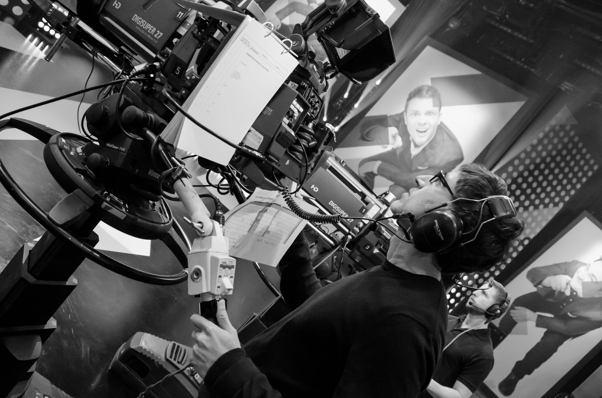 A man operating camera equipment on a set, with a large screen behind him displaying a man in a suit.