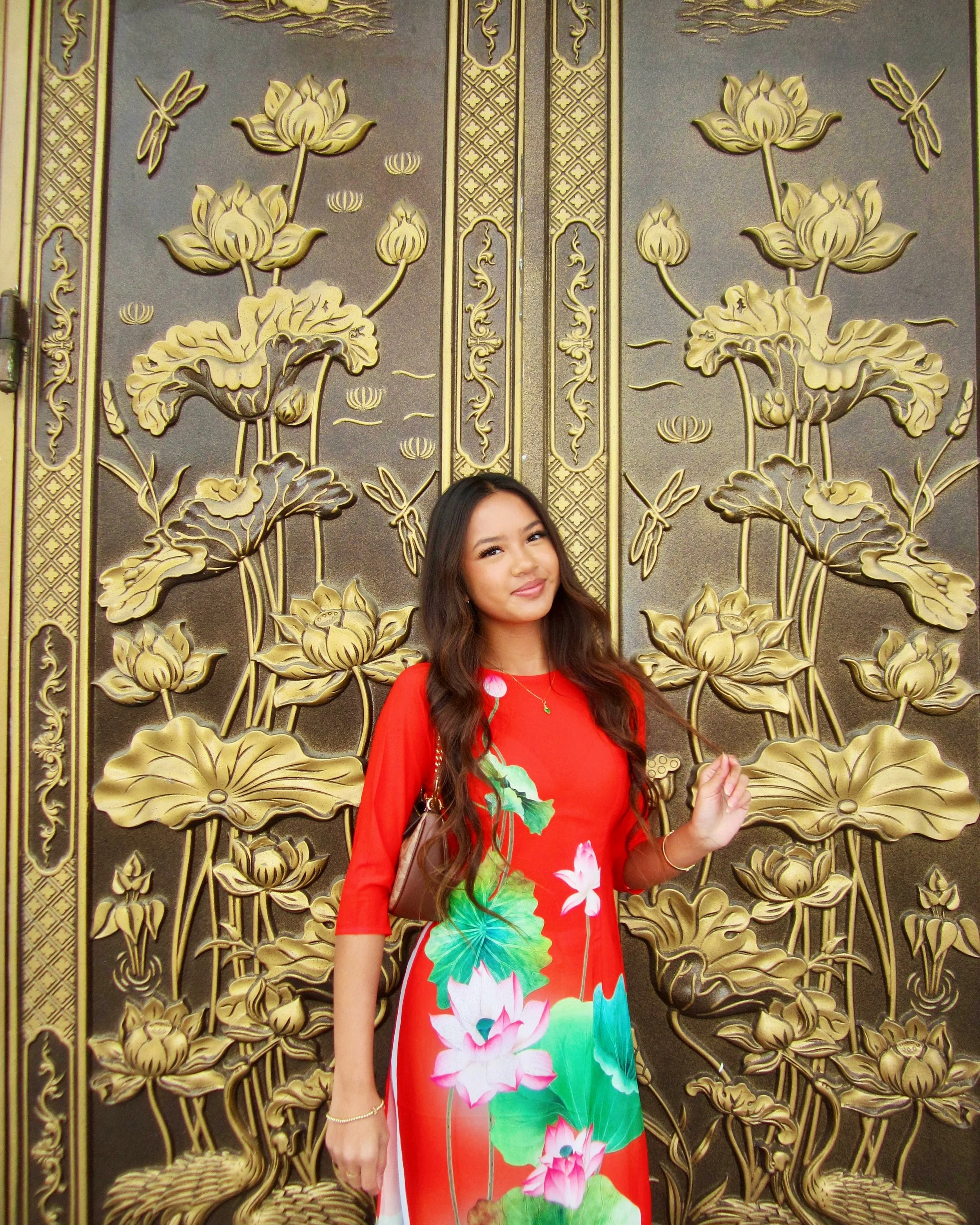 A woman in a red dress with floral patterns stands in front of a decorative golden door with lotus flower and dragonfly motifs.
