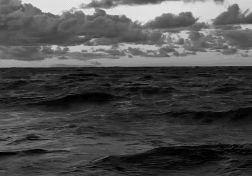Black and white photo of a cloudy sky over the ocean with waves in the foreground.