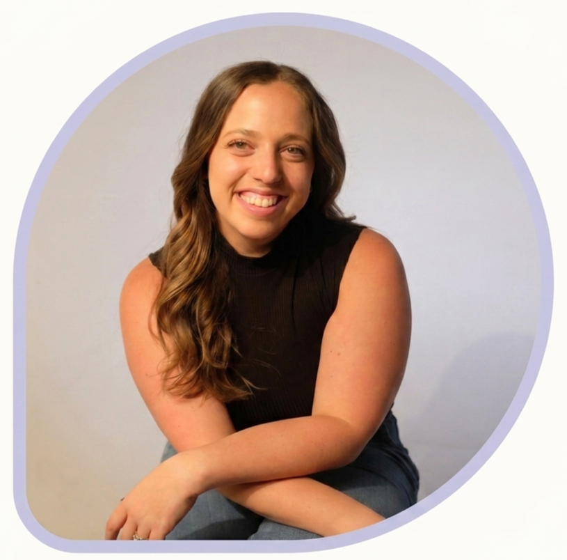 A young woman with long brown hair, smiling, wearing a black sleeveless top, sitting against a plain light-colored background.