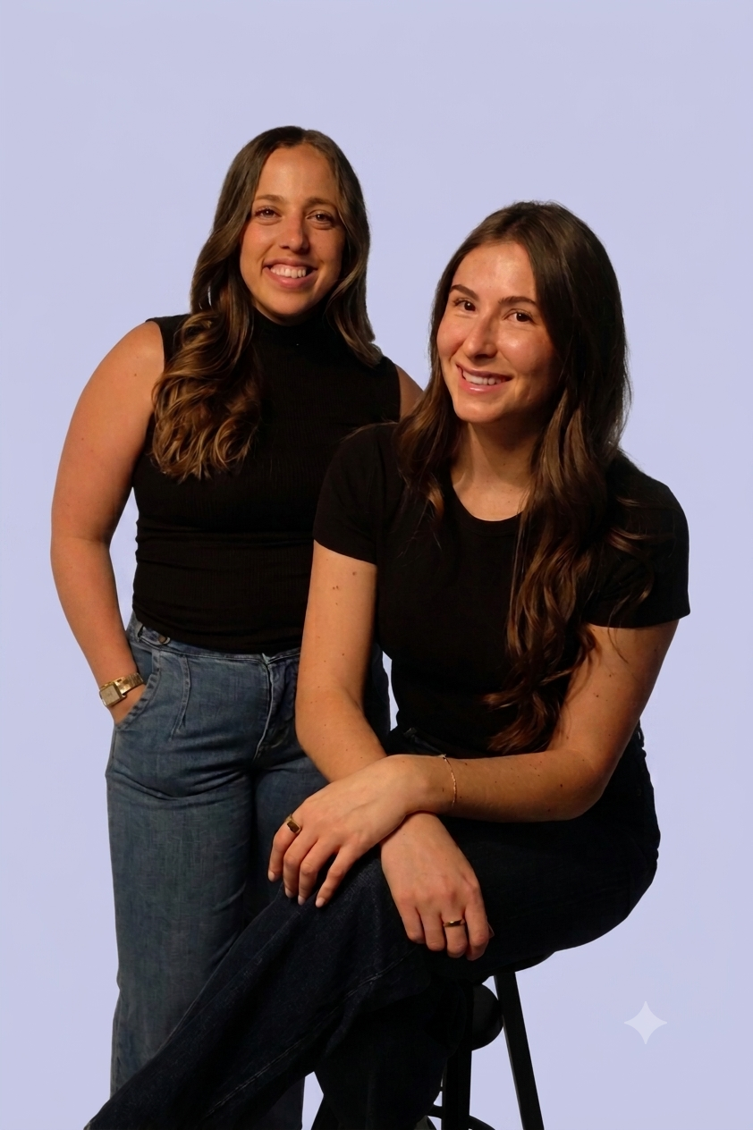 Two women smiling, one standing and one sitting on a stool, both wearing black tops and jeans, against a light purple background.