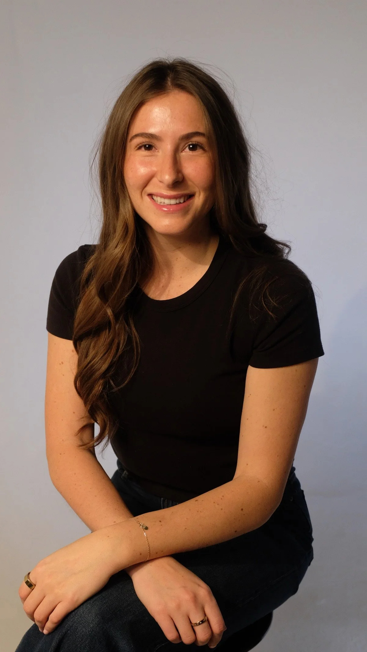 A young woman with long wavy brown hair, wearing a black t-shirt and blue jeans, sitting against a light blue background, smiling at the camera.