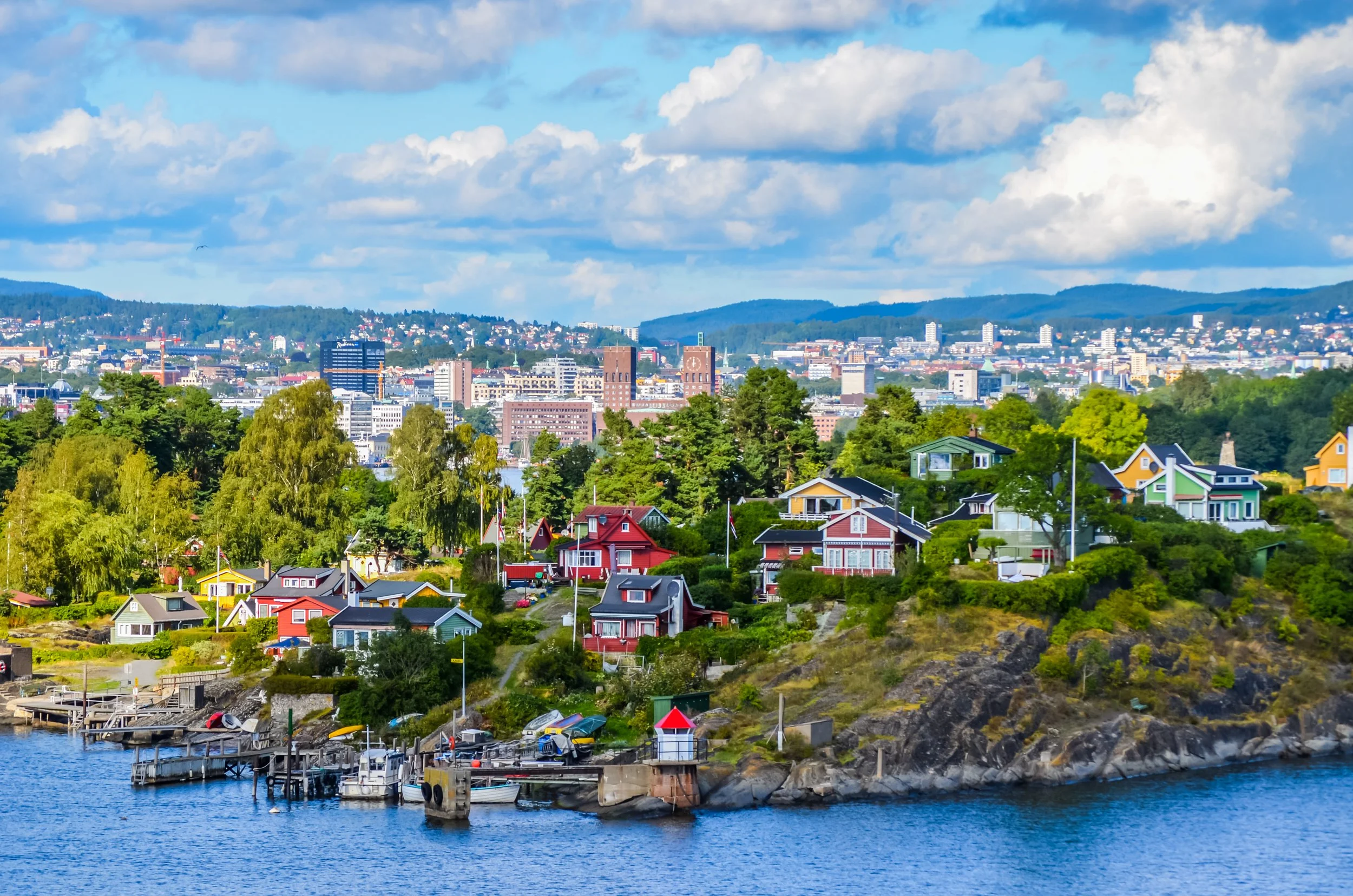 En kuststad med färgglada hus och en pir vid vattnet. I bakgrunden syns stadens högre byggnader och gröna kullar under en molnig himmel.