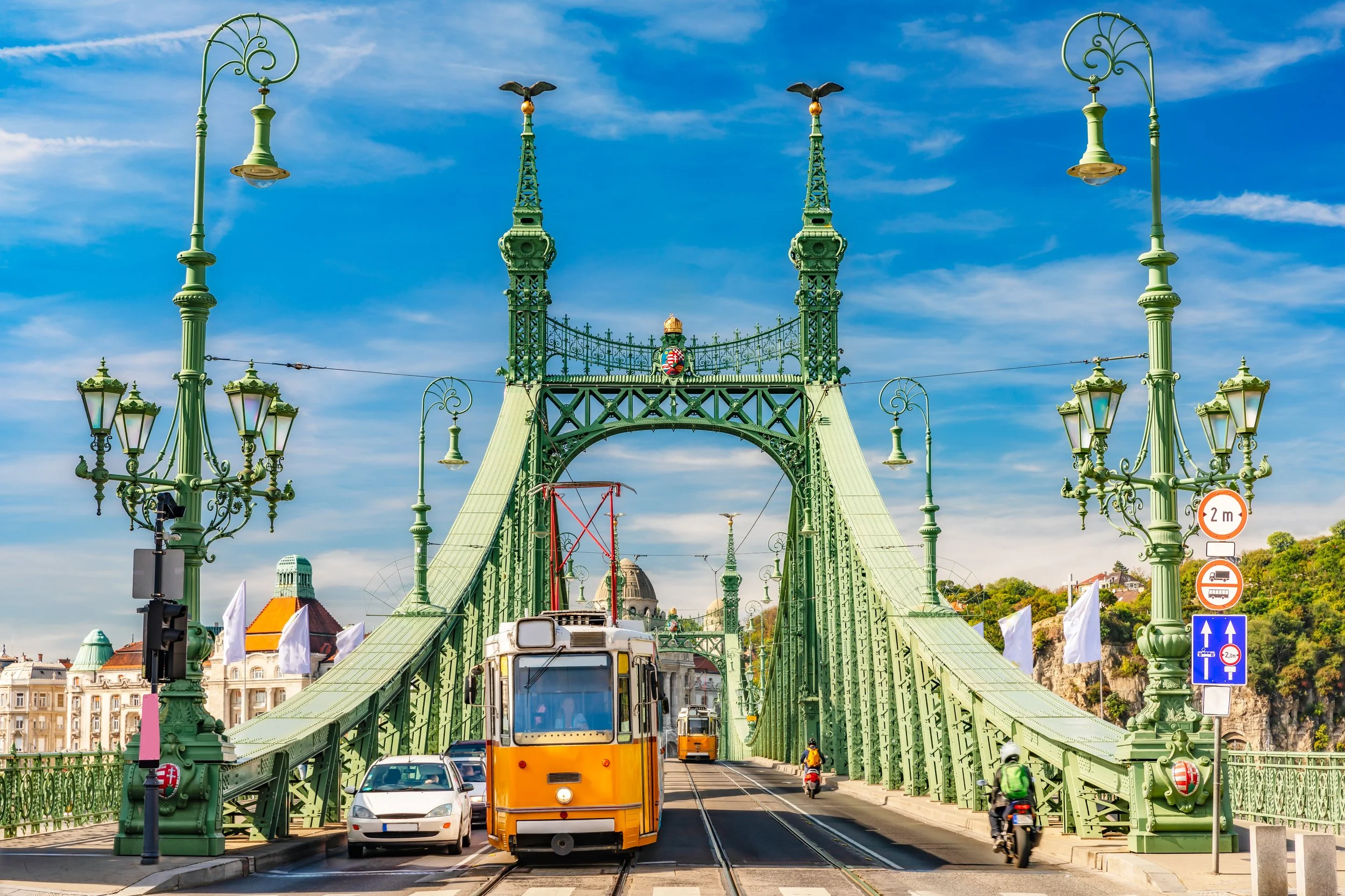 Vy av Széchenyi Lánchíd i Budapest med tåg, bilar och motorcyklar på bron, samt lampor och dekorativa element på järnbron, mot en blå himmel.