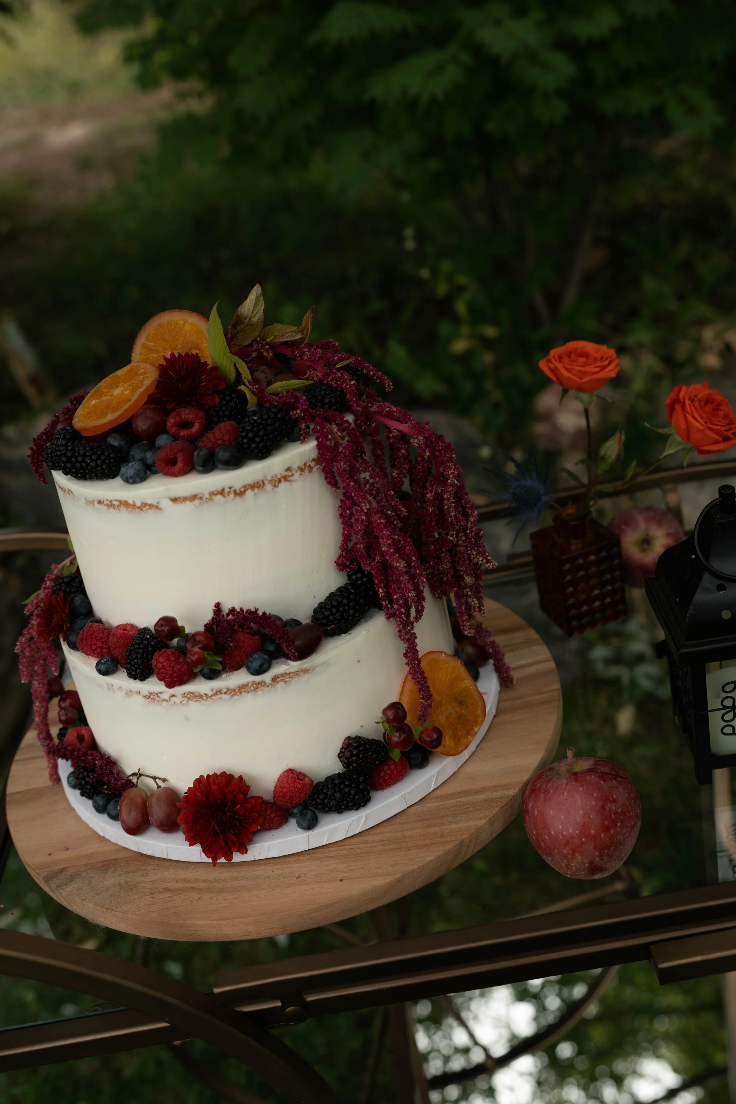 Two-tier white naked cake decorated with berries, orange slices, dark flowers, and cascading burgundy blooms on a wooden dessert table, with orange roses in the background.