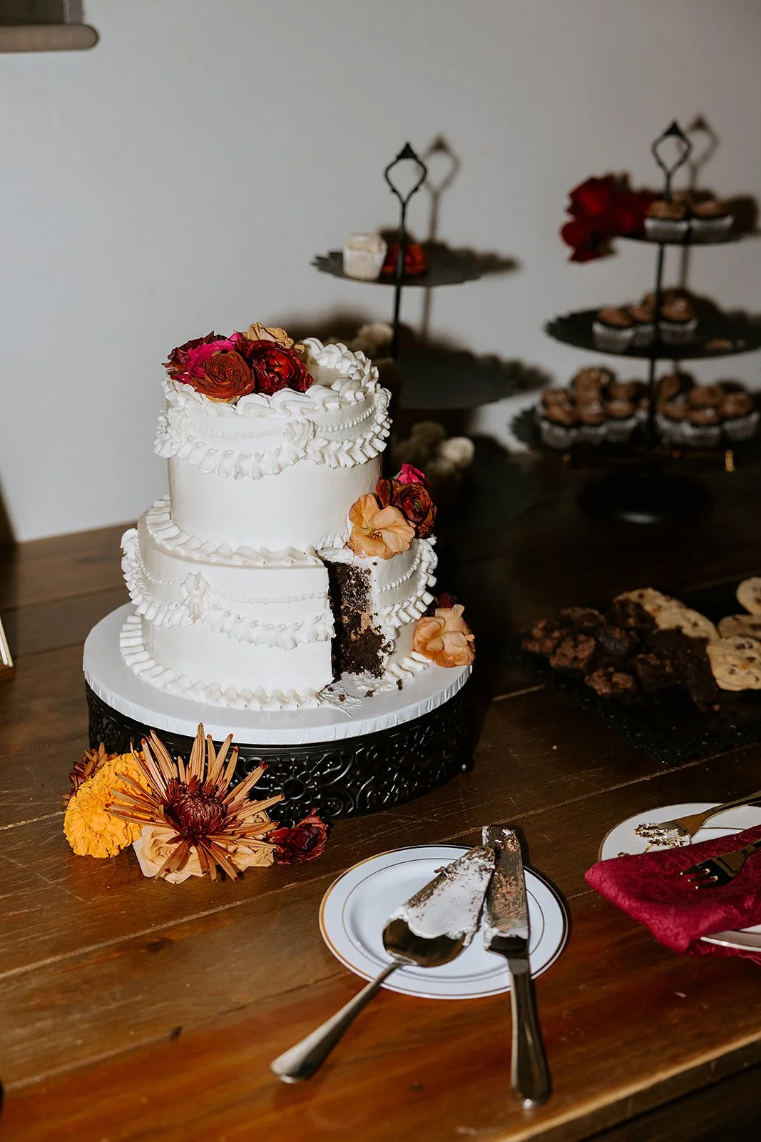 Three-tier white wedding cake decorated with dried flowers, a slice removed, dessert plates, and pastries on a wooden table.