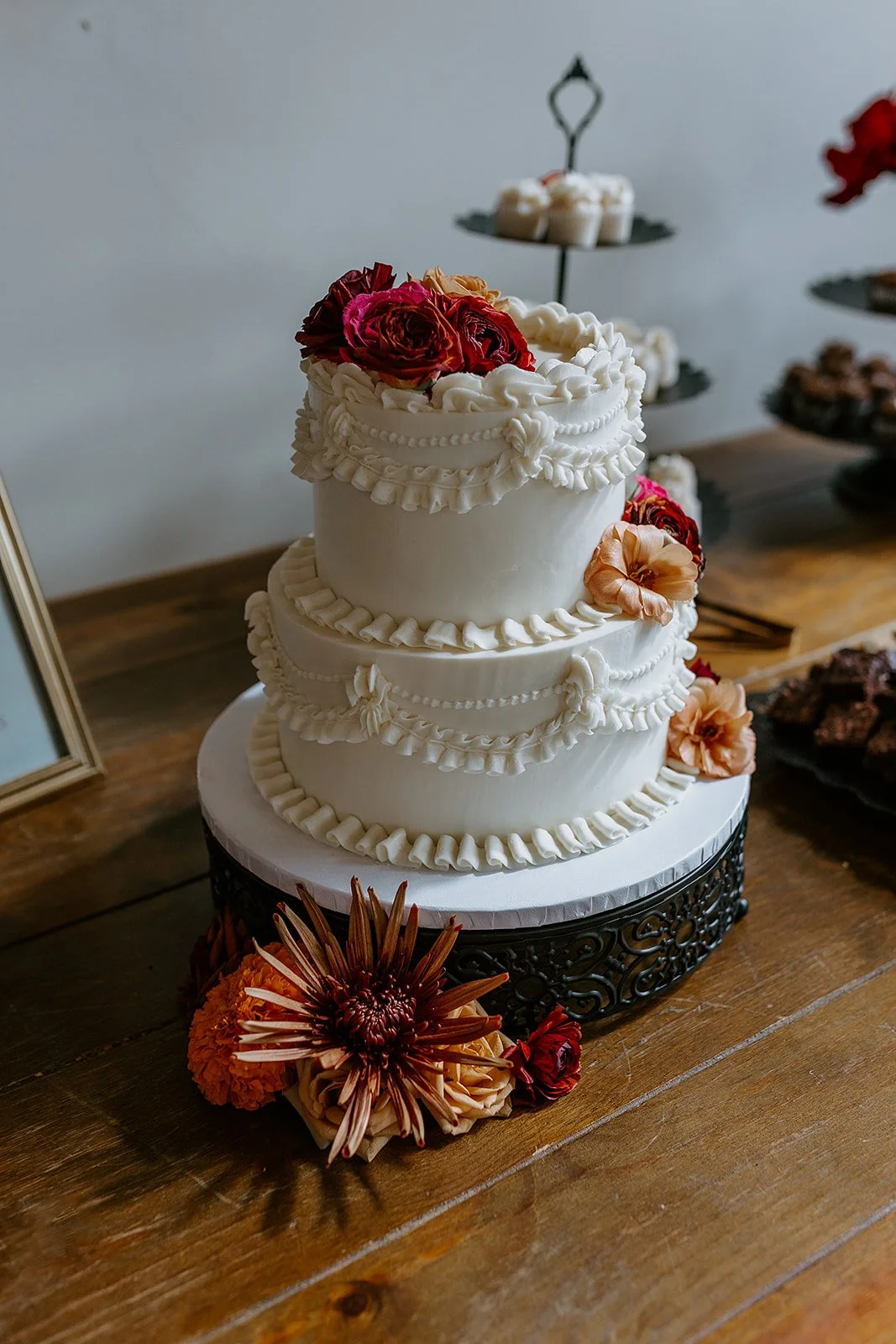 Three-tier white wedding cake with ornate buttercream piping and red, peach, and orange flowers on a wooden table