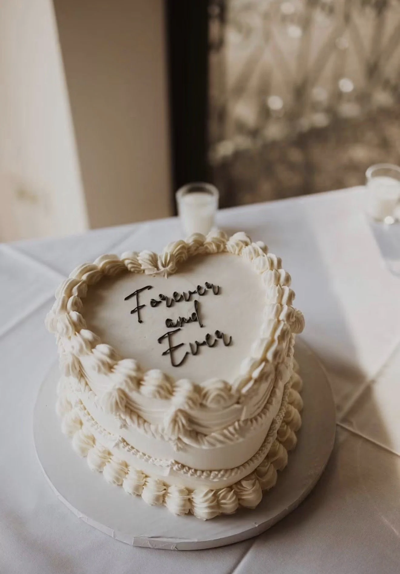 Ornate white celebration cake with piped frosting and chocolate lettering reading “Forever and Ever” on a table.
