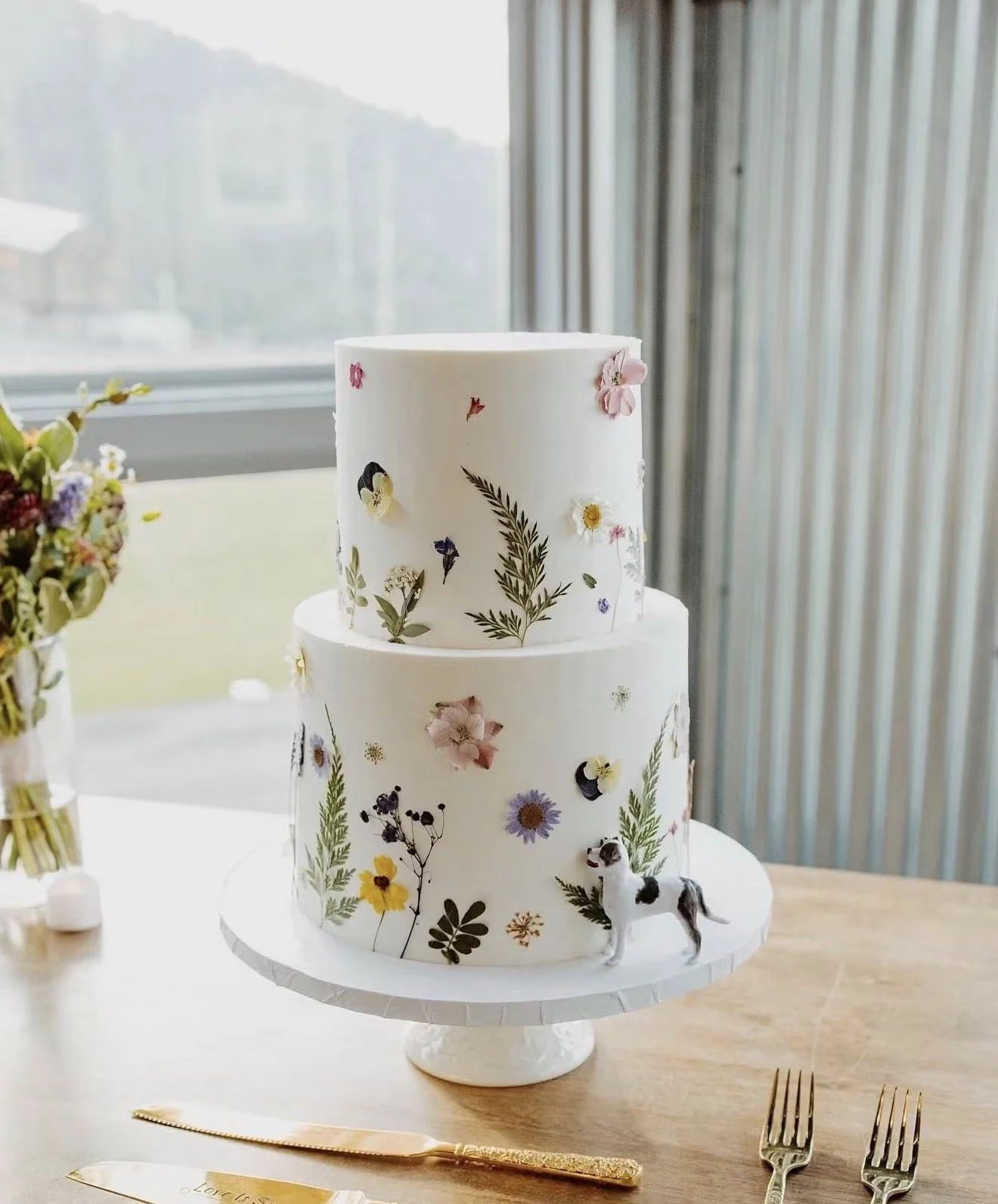 Two-tier white wedding cake decorated with pressed flowers, greenery, and a small black-and-white dog figurine on a cake stand