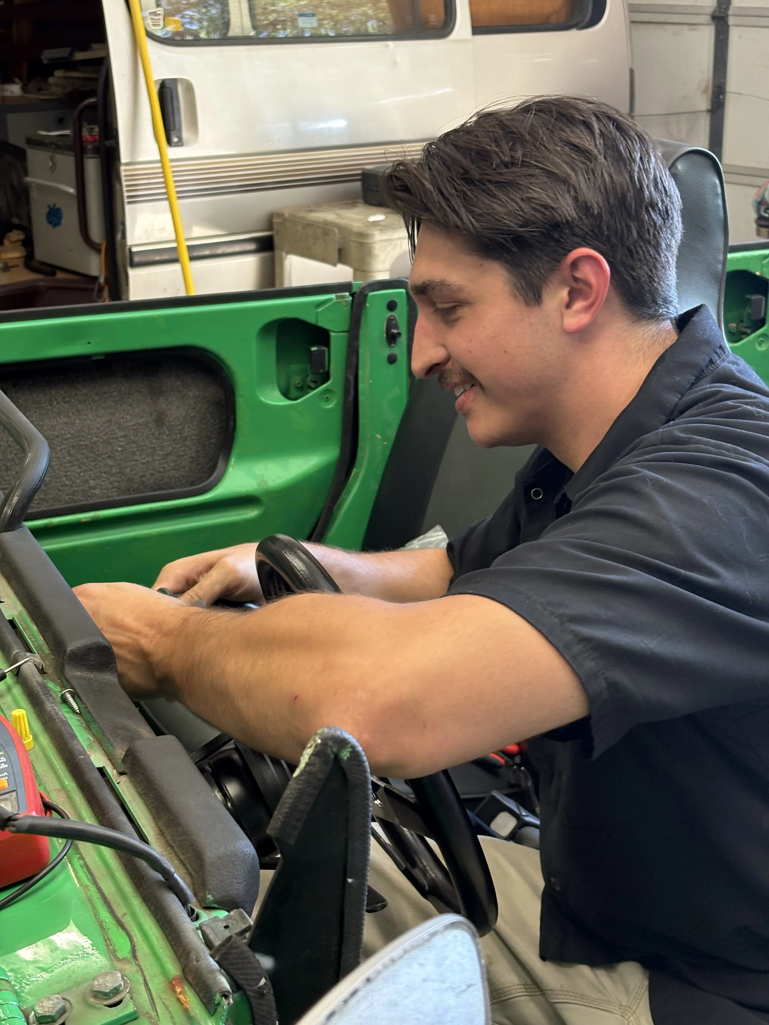 A mechanic with brown hair, wearing a black shirt, is working on a green vehicle in a garage or workshop. He is smiling and focused on his task.