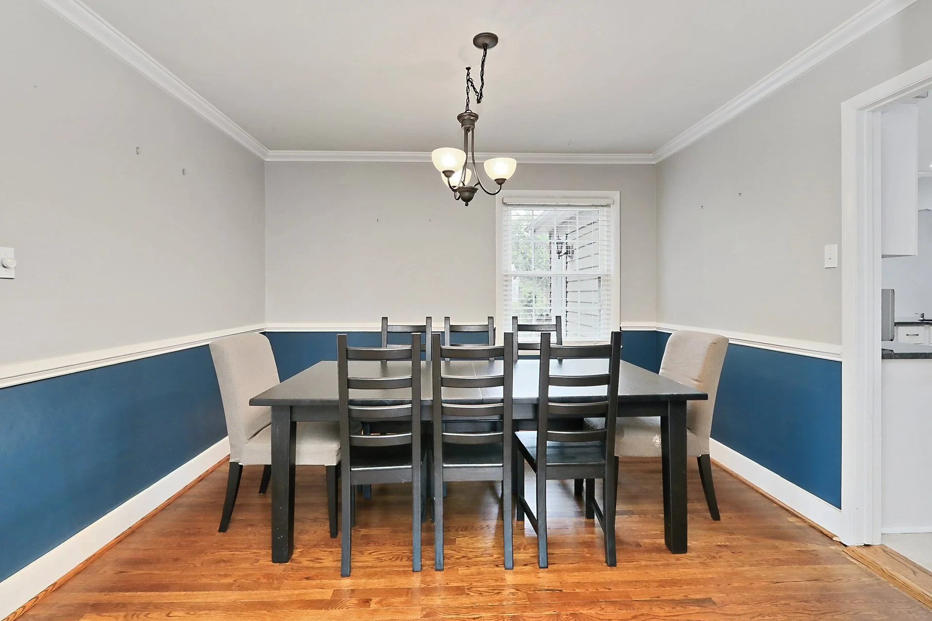 Empty dining room with black table, eight chairs, two beige armchairs, window, and blue wainscoting.