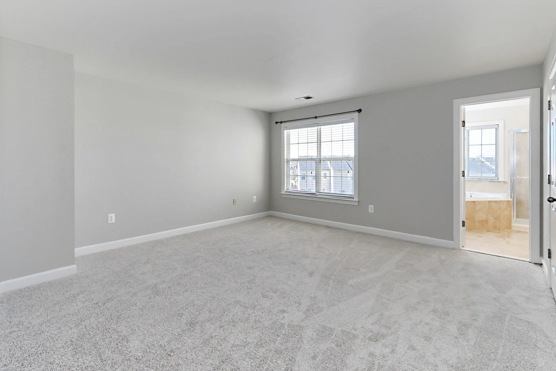 Empty living room with large window and doorway leading to bathroom, beige carpeted floor, white walls, bright natural light.