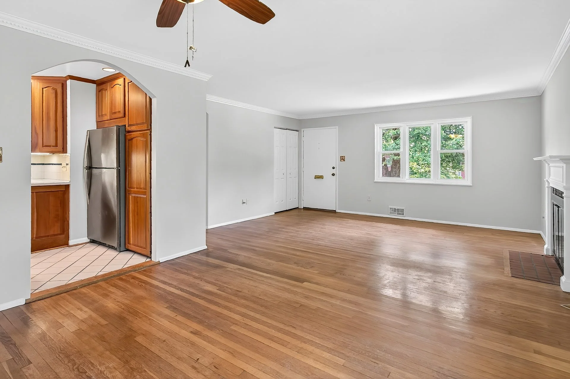 Empty living room with hardwood floors, white walls, large window, and a fireplace, with an open kitchen area visible to the left.