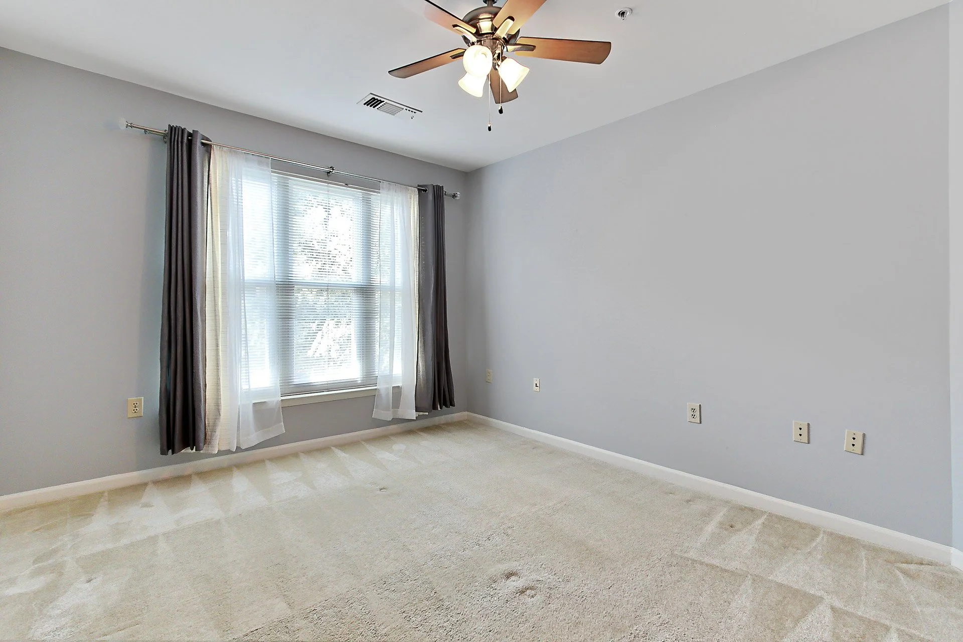 Empty bedroom with light gray walls, beige carpet, window with sheer white and dark gray curtains, ceiling fan with light, and electrical outlets on the walls.