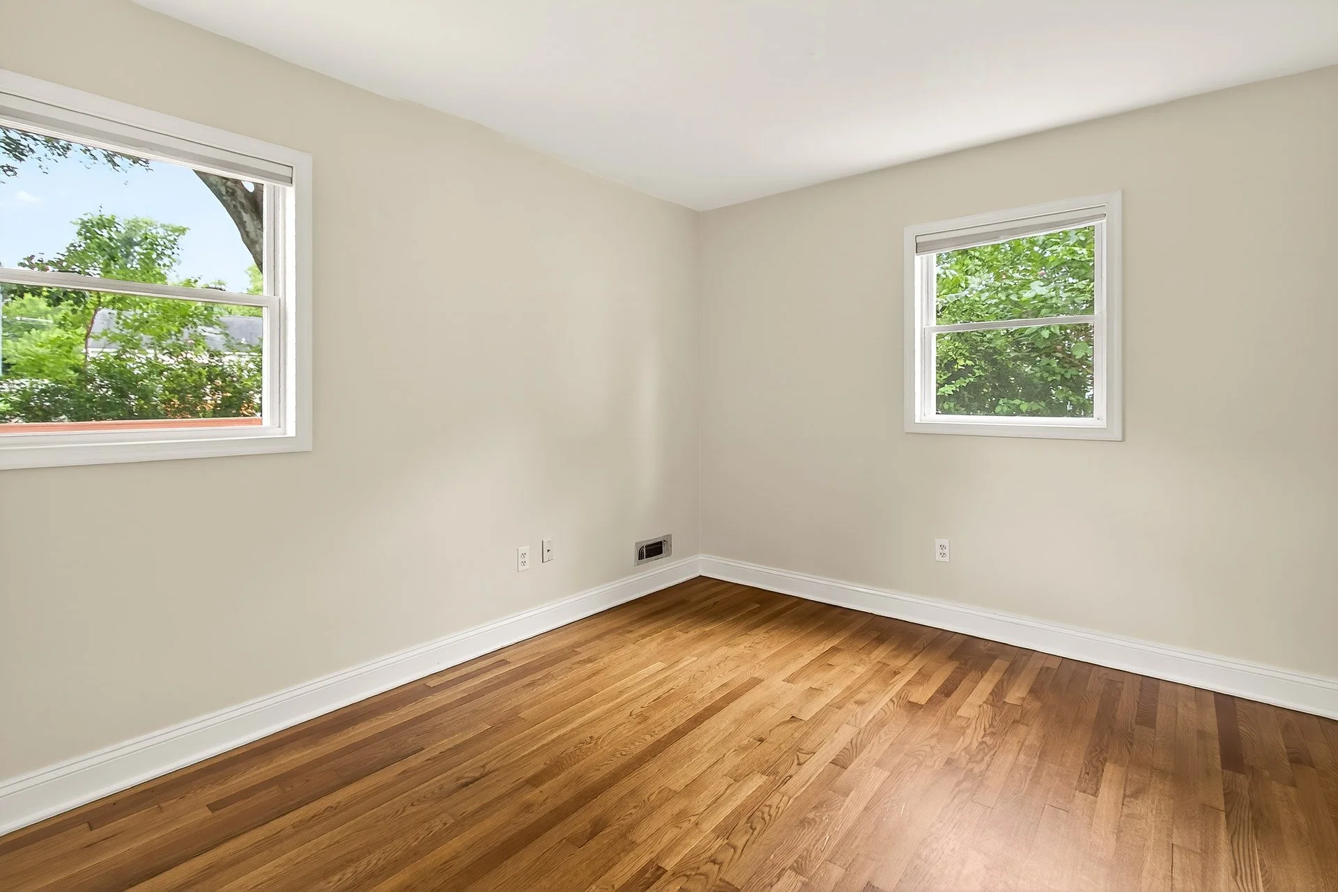 Empty room with hardwood floors, two windows with white trim, and beige walls.
