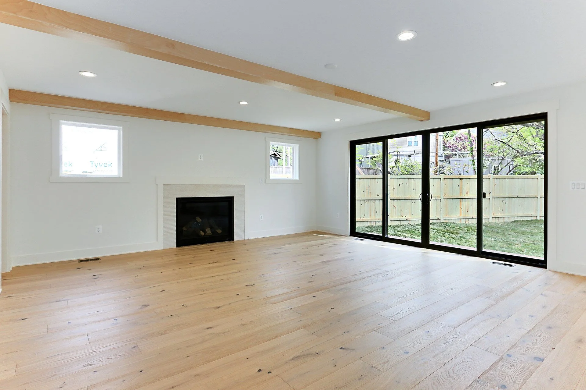 Empty living room with light wood flooring, white walls, small windows, a fireplace, and large sliding glass doors leading to a backyard with a wooden fence and trees.