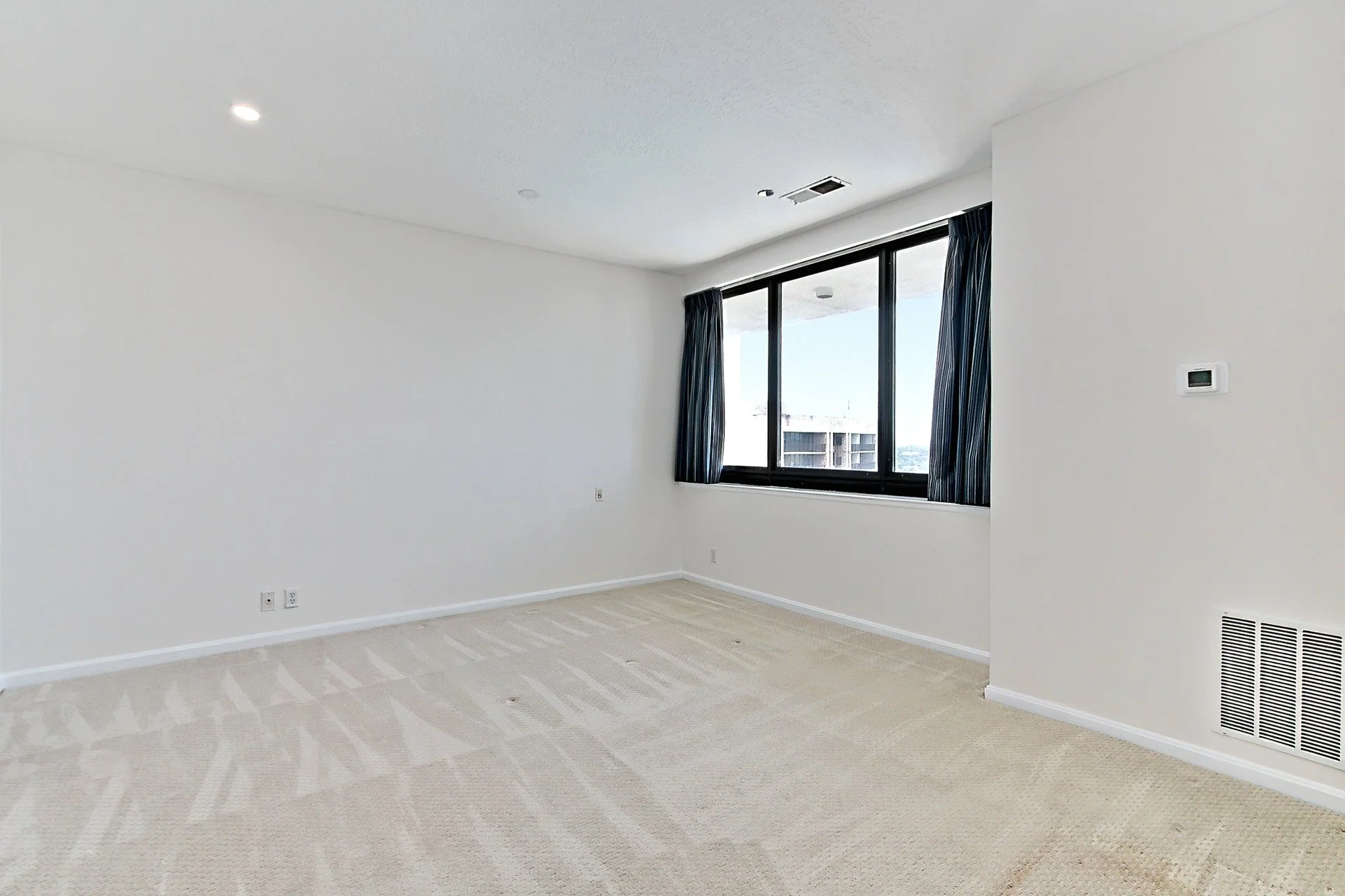Empty room with beige carpet, white walls, and large window with dark curtains.