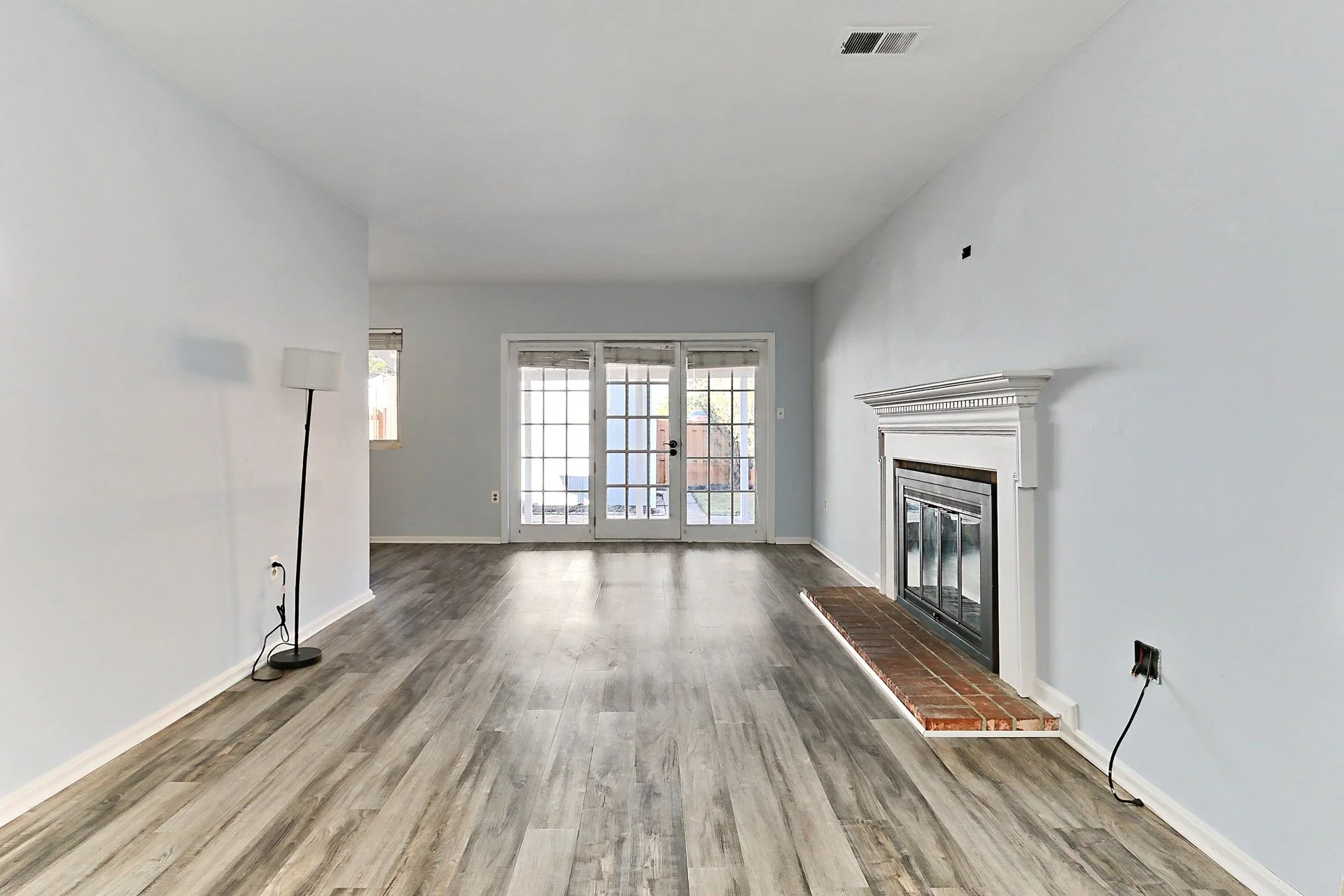 Empty living room with gray wood floors, white walls, a fireplace with brick hearth, and double glass doors leading outside.