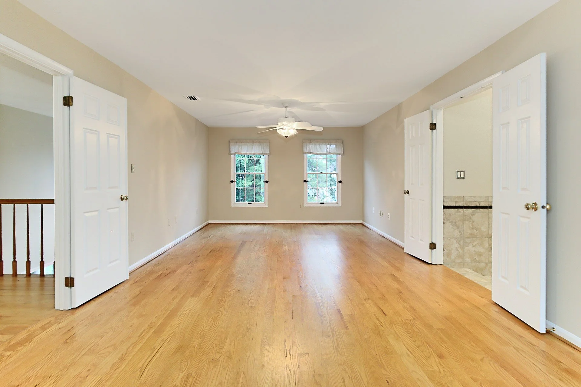 Empty living room with hardwood floors, beige walls, two windows with curtains, a ceiling fan, and two open doorways.