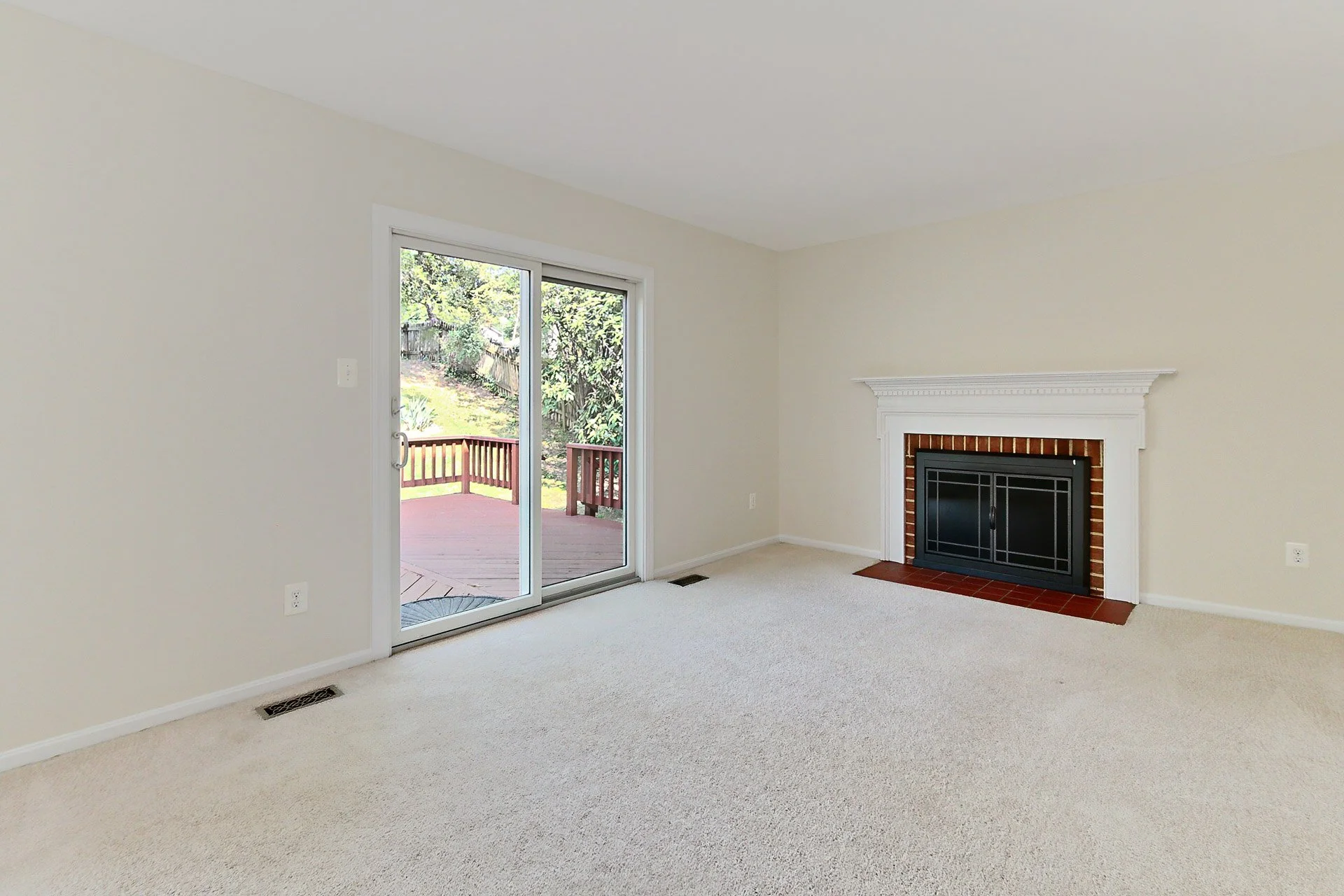 Empty living room with a fireplace, sliding glass door leading to a wooden deck, beige carpet, white walls, and a view of a backyard with greenery.