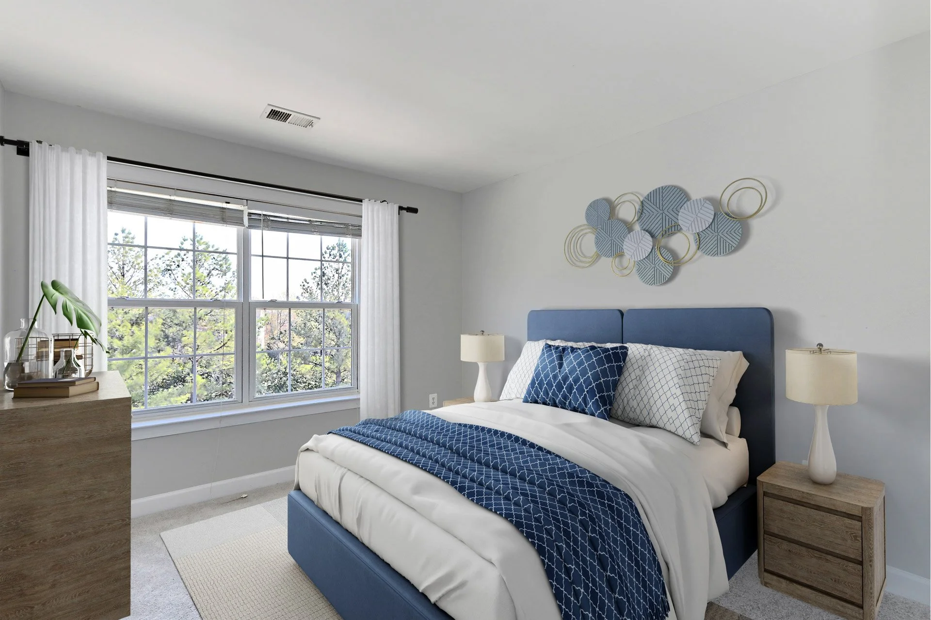 Modern bedroom with large window, blue upholstered bed, white and blue bedding, wooden nightstand with a lamp, abstract wall art, and a wooden dresser with decorative items.