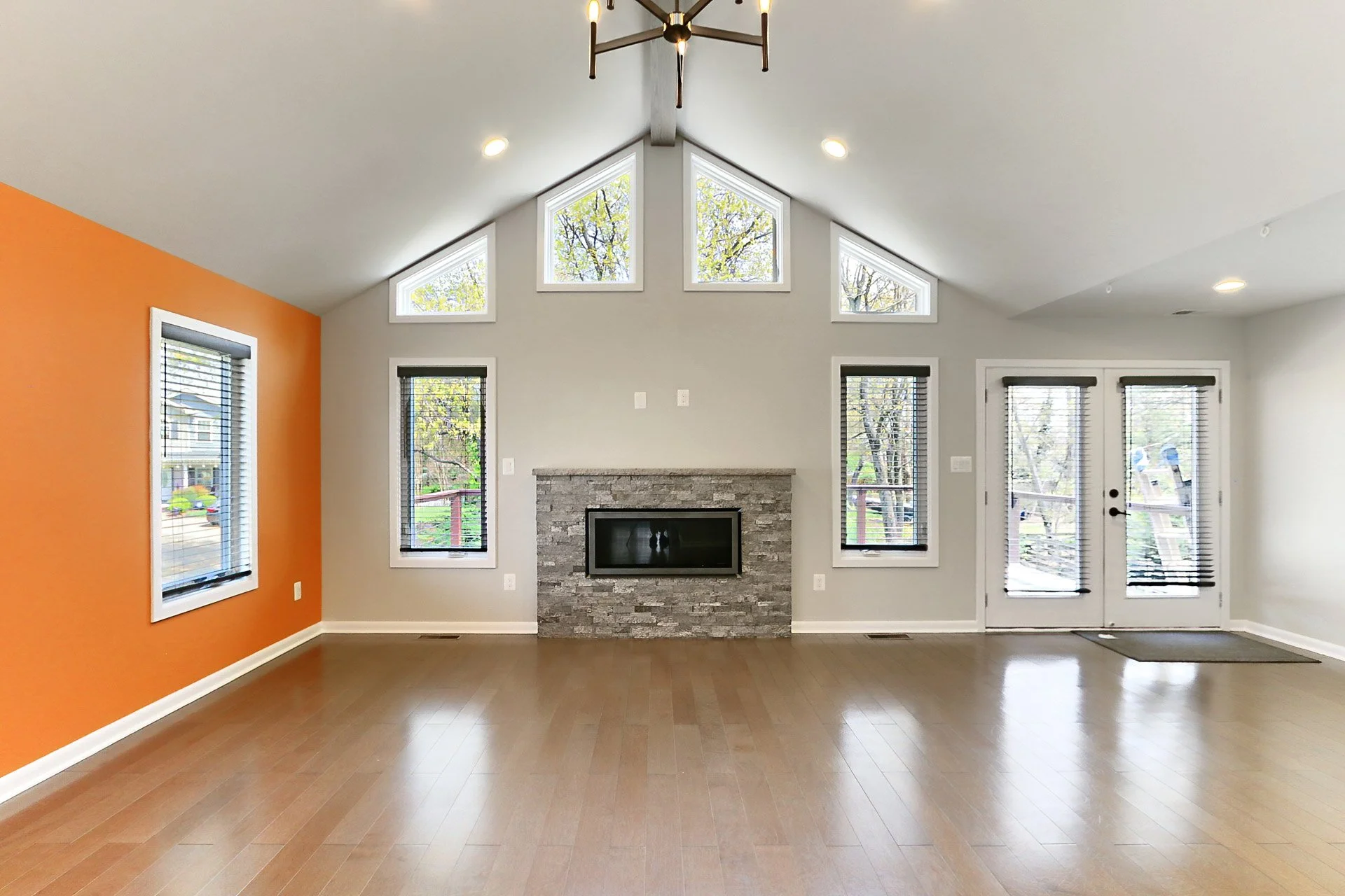 Empty living room with large windows, a stone fireplace, and French doors, with an orange accent wall and hardwood floors.