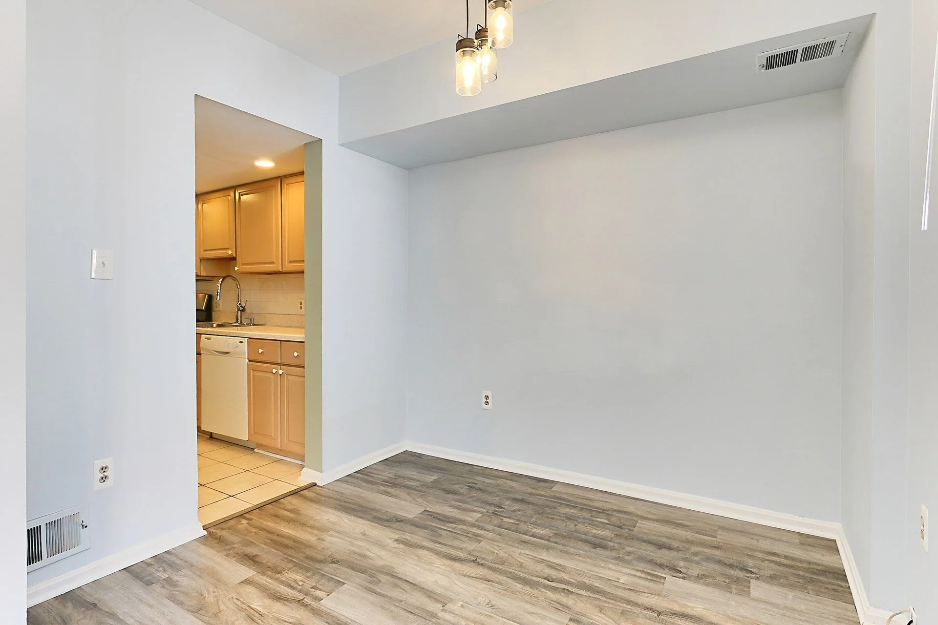 Empty living room with white walls, wood laminate floors, a small section of kitchen with beige cabinets visible through a doorway, ceiling light fixture, and air vent.