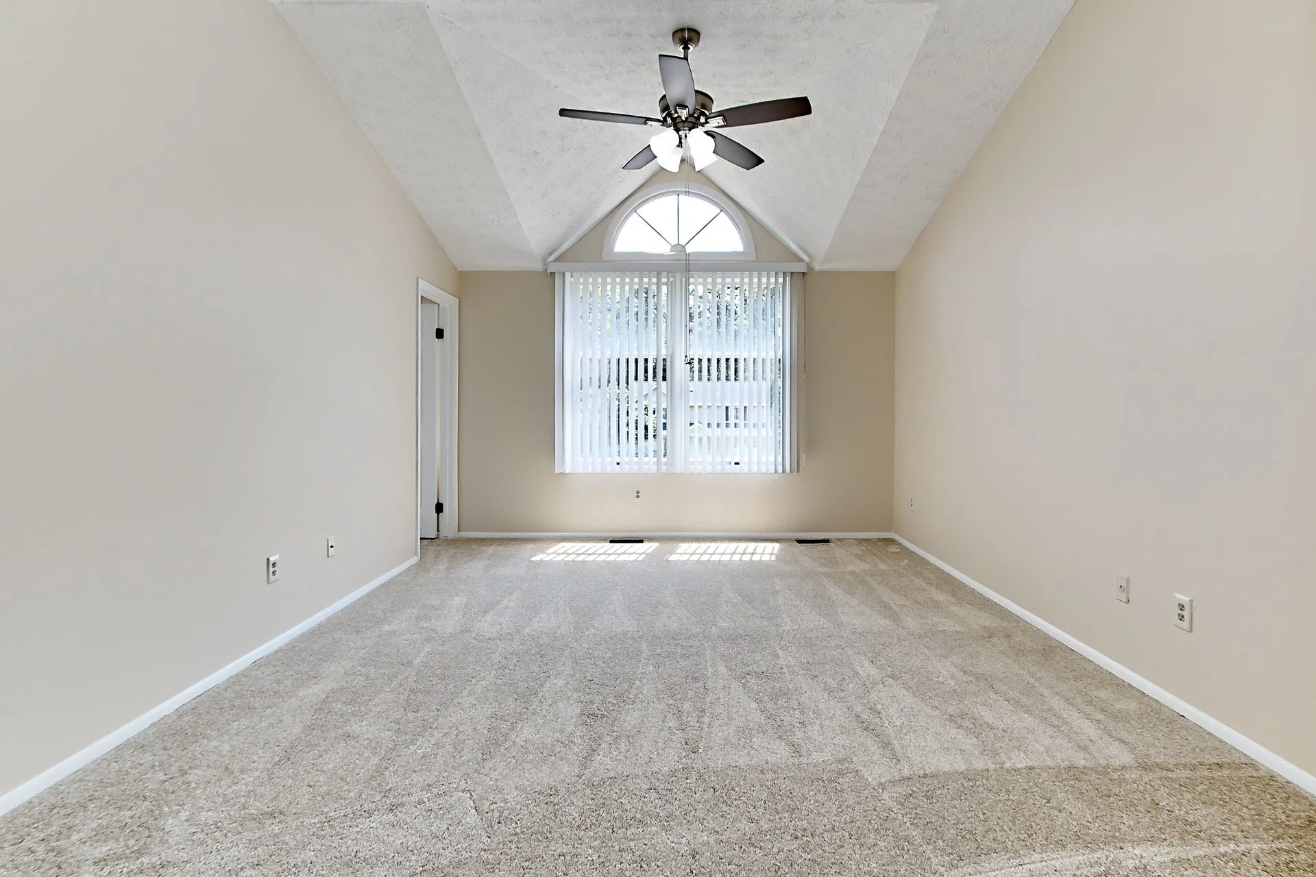 Empty room with beige walls, carpeted flooring, a ceiling fan, a large window with vertical blinds, and a door on the left