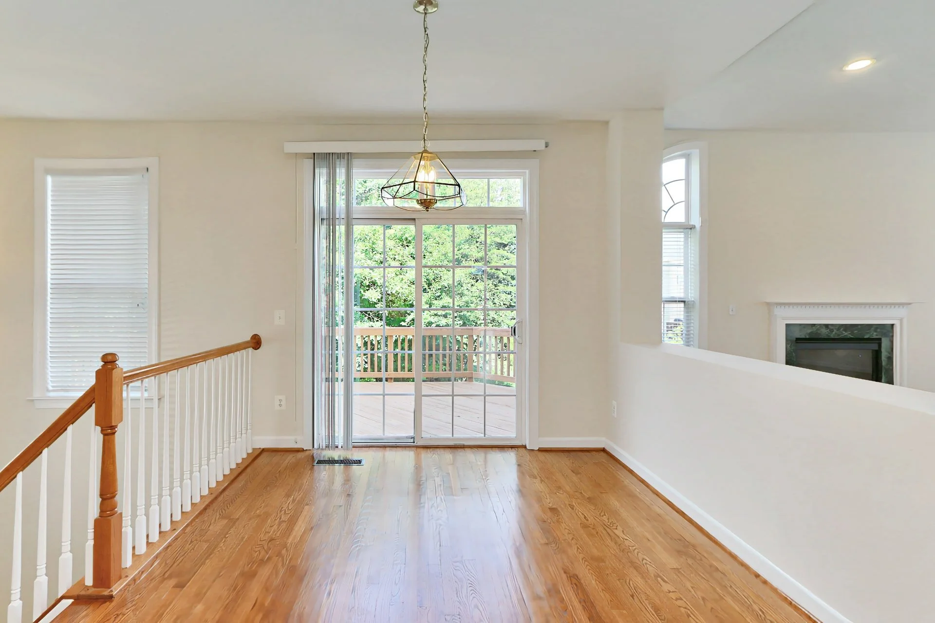 Interior view of a bright living room with hardwood floors, a sliding glass door leading to a wooden porch outside, and windows with blinds. There's a decorative hanging lamp.