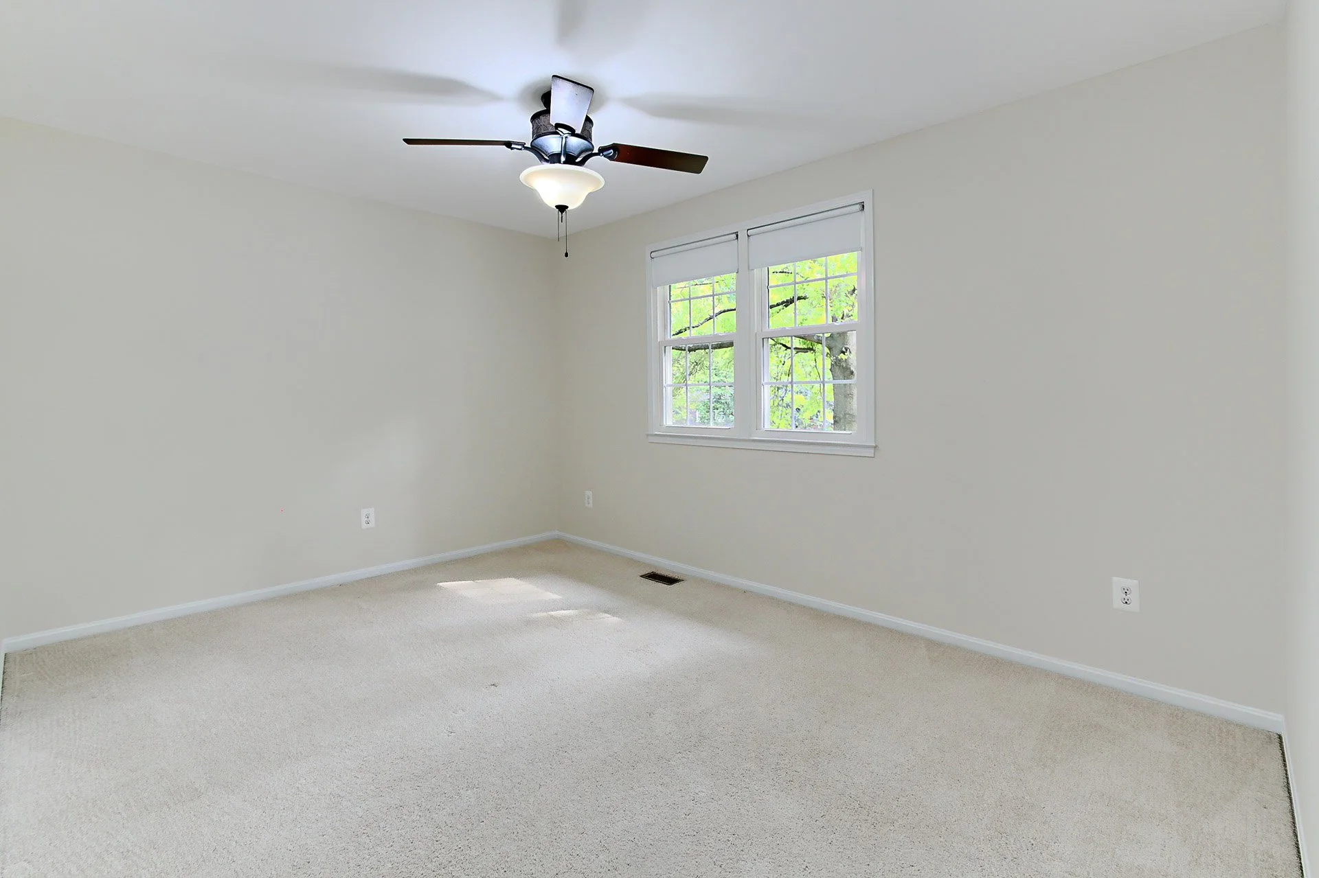 Empty bedroom with beige carpet, white walls, a ceiling fan with light, and a window showing green trees outside.