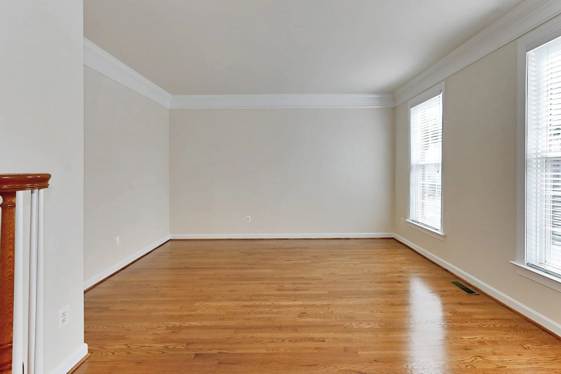 Empty living room with hardwood floors, beige walls, white crown molding, and three large windows with blinds.
