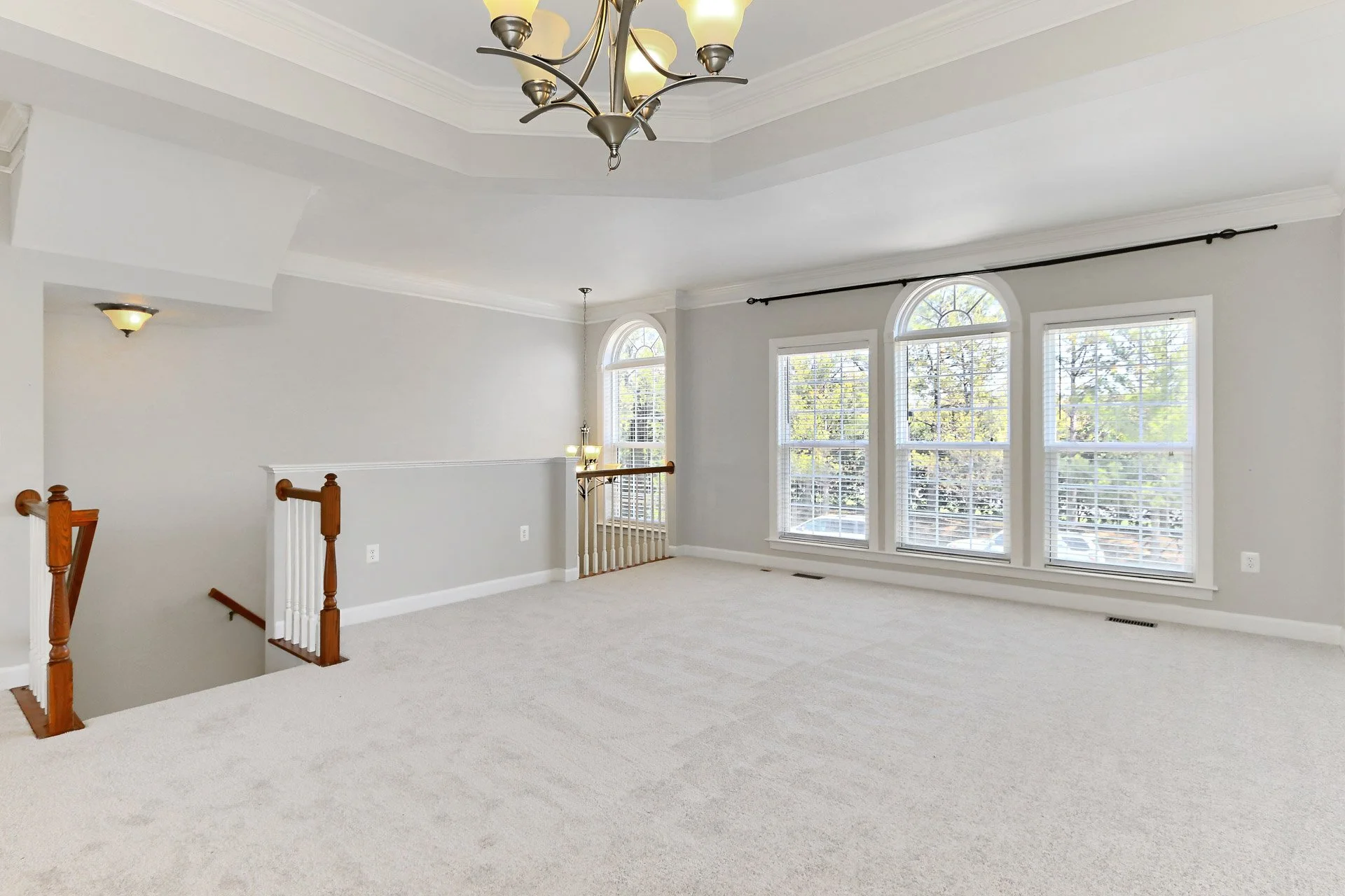 An empty living room with white walls, large windows with blinds, a chandelier hanging from the ceiling, and a staircase with wooden banisters leading downstairs.