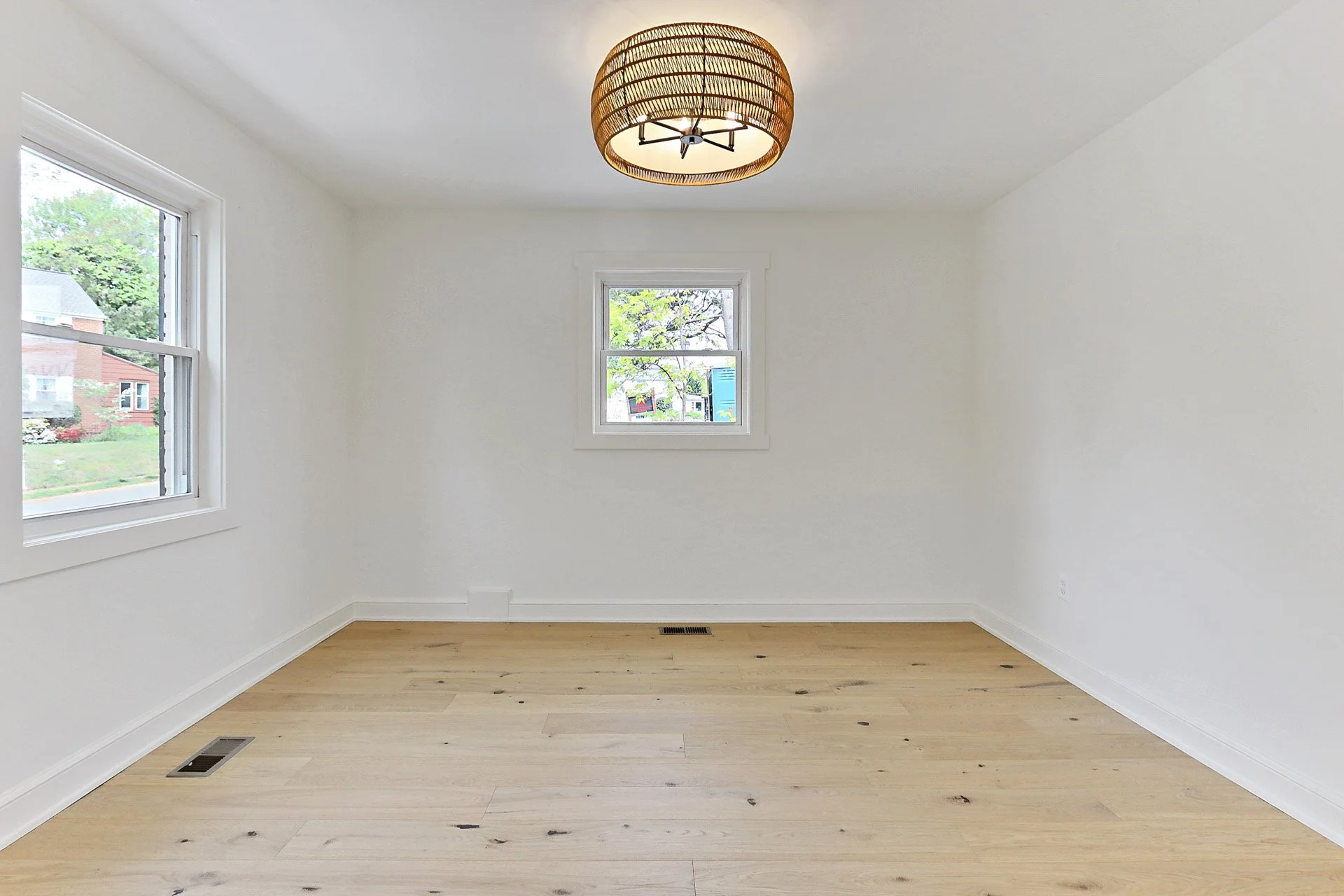 Empty room with white walls, light wooden flooring, two windows, and a ceiling light fixture.